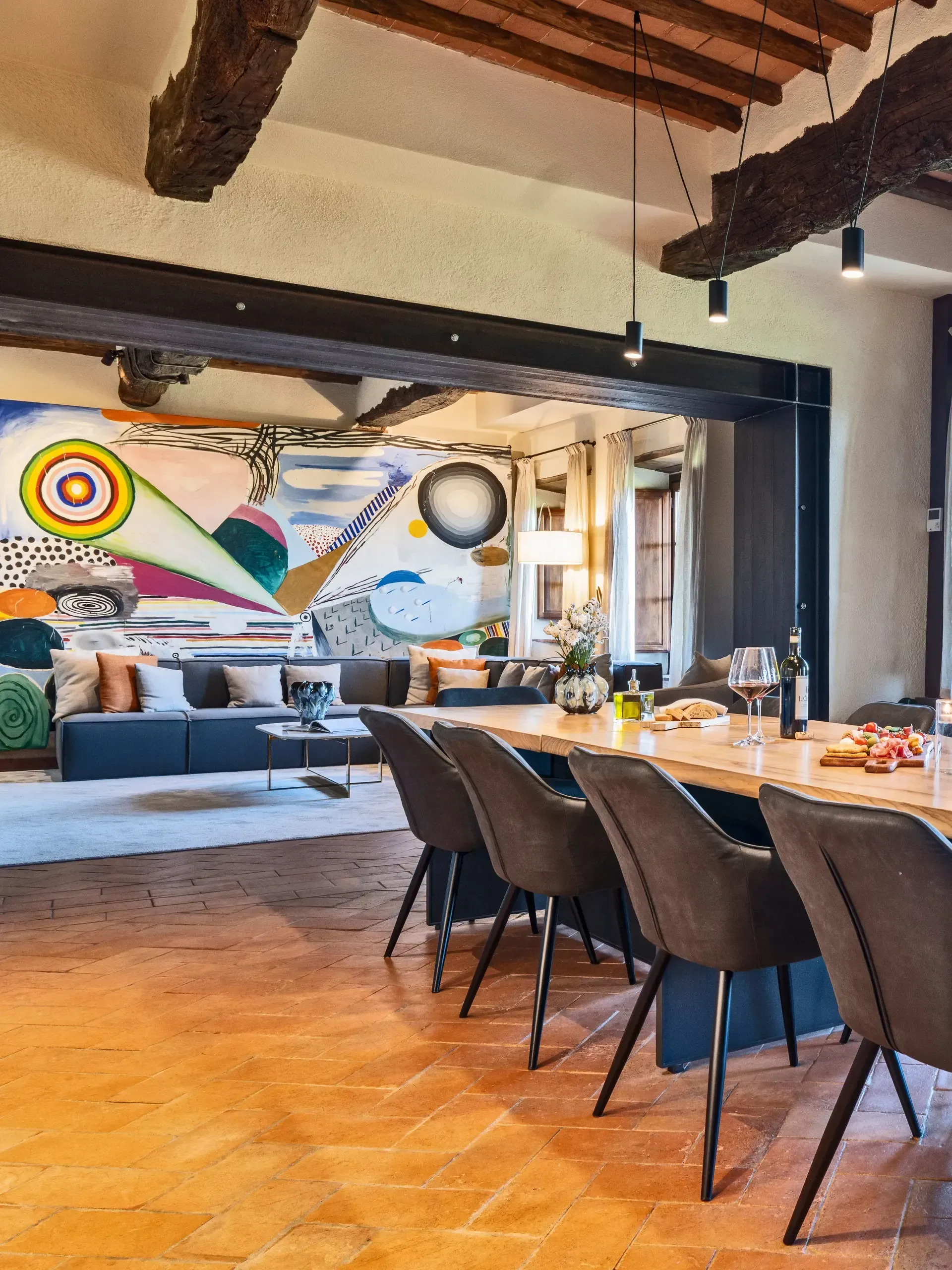 Dining area with long table and chairs; colorful mural in the background, exposed wooden beams.