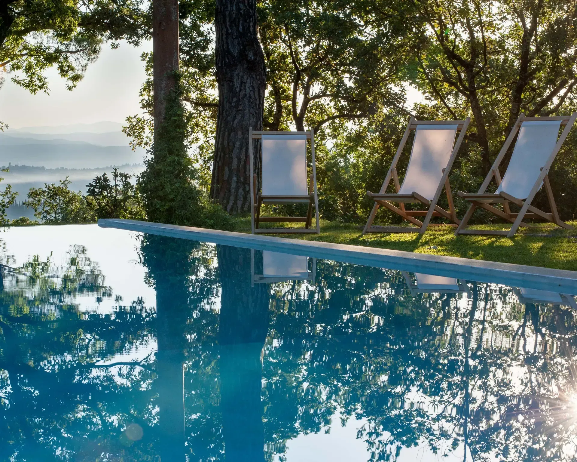 Poolside scene with a long rectangular pool reflecting trees and sky, three white-and-wood deck chairs on the grass with a view of rolling hills.
