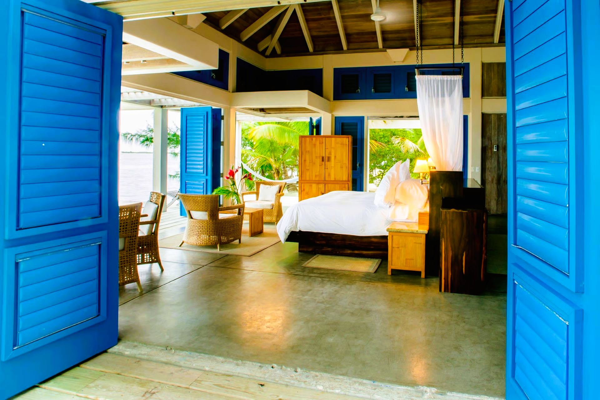 Blue-shuttered doorway opens to a spacious bedroom with a bed, furniture, and view of greenery.