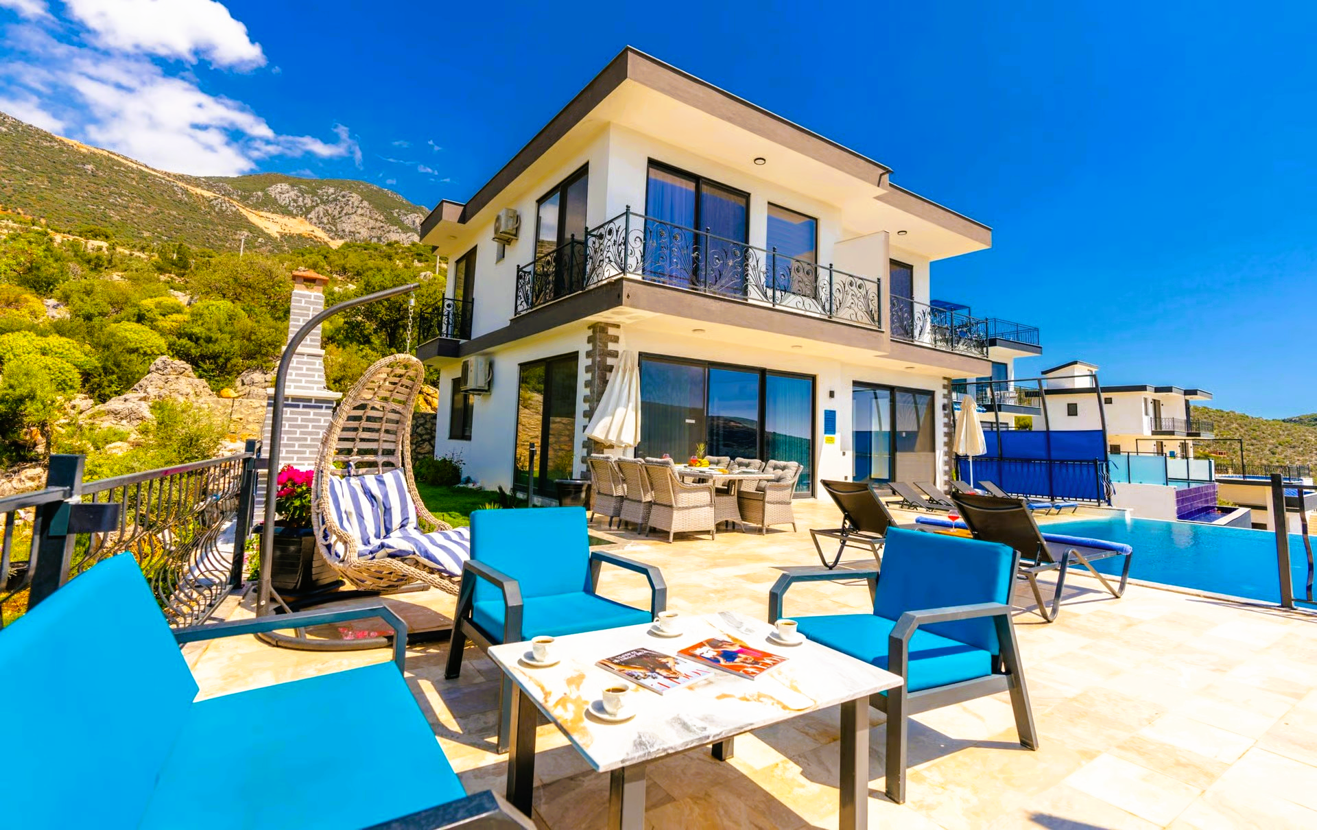 Modern villa with blue accents, patio furniture, pool, and mountain backdrop under a sunny sky.