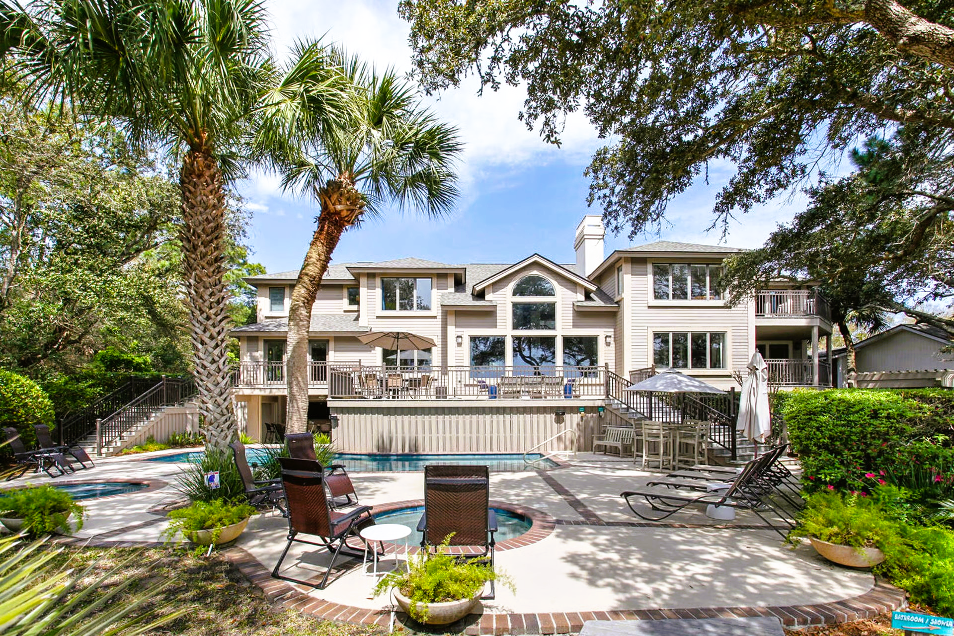 Backyard with pool, spa, patio furniture, and multi-story beige house under sunny sky.