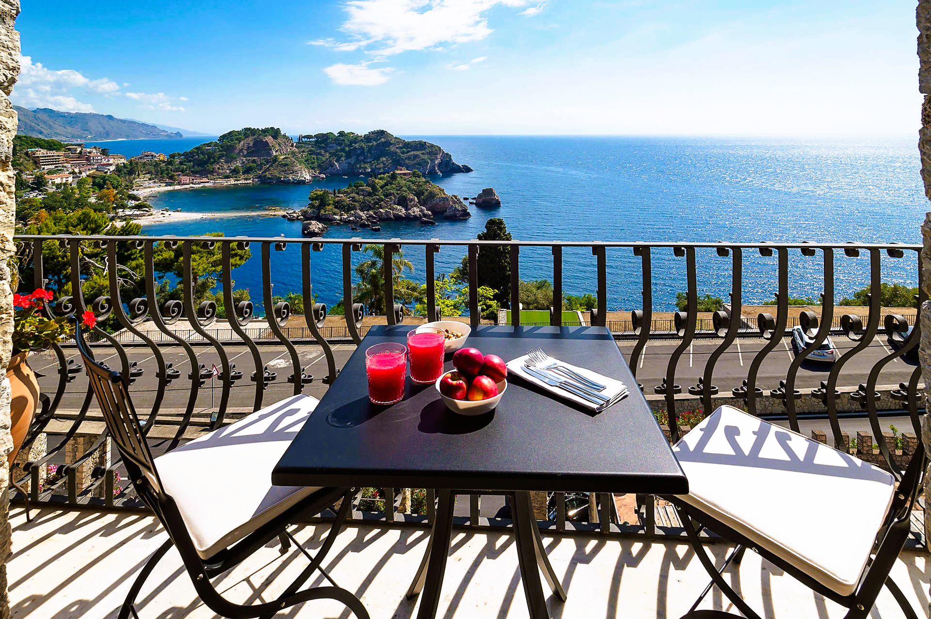 Balcony overlooking the sea; table with drinks and fruit, two chairs. Blue water, sunny day.