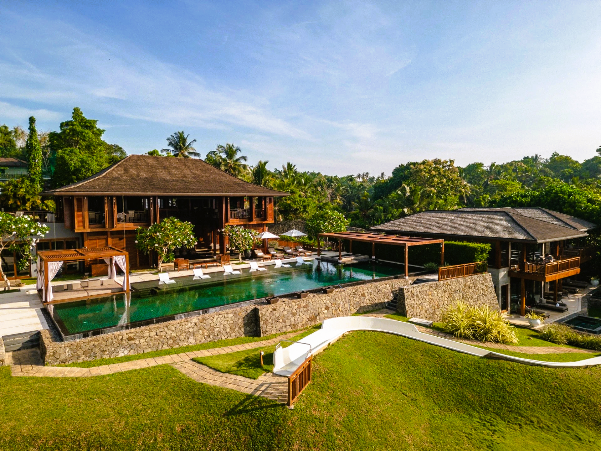 Luxurious wooden villa with pool, lush greenery, and blue sky, overlooking a tropical landscape.