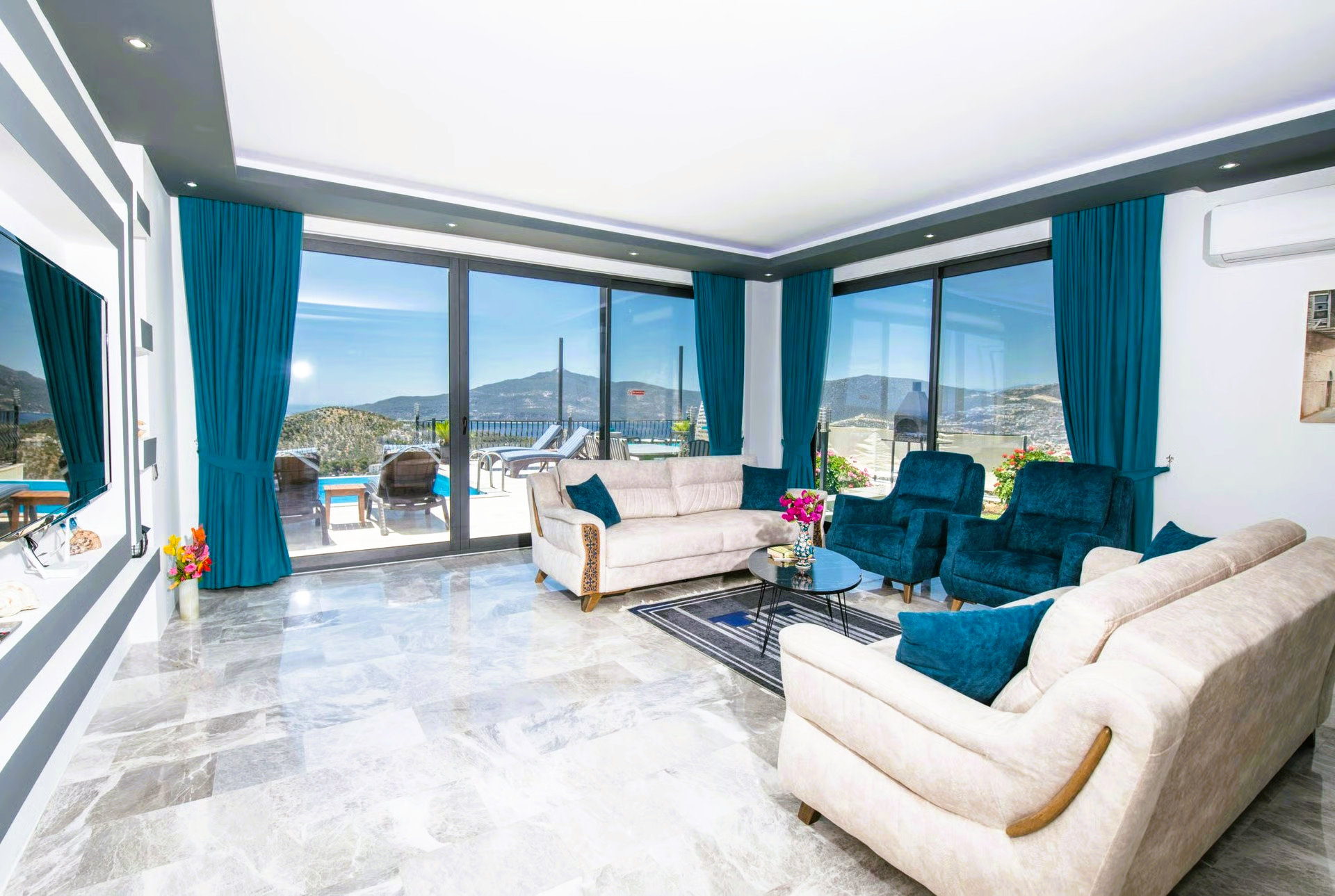 Living room with large windows, light-colored sofas, teal curtains, overlooking a pool and mountains.