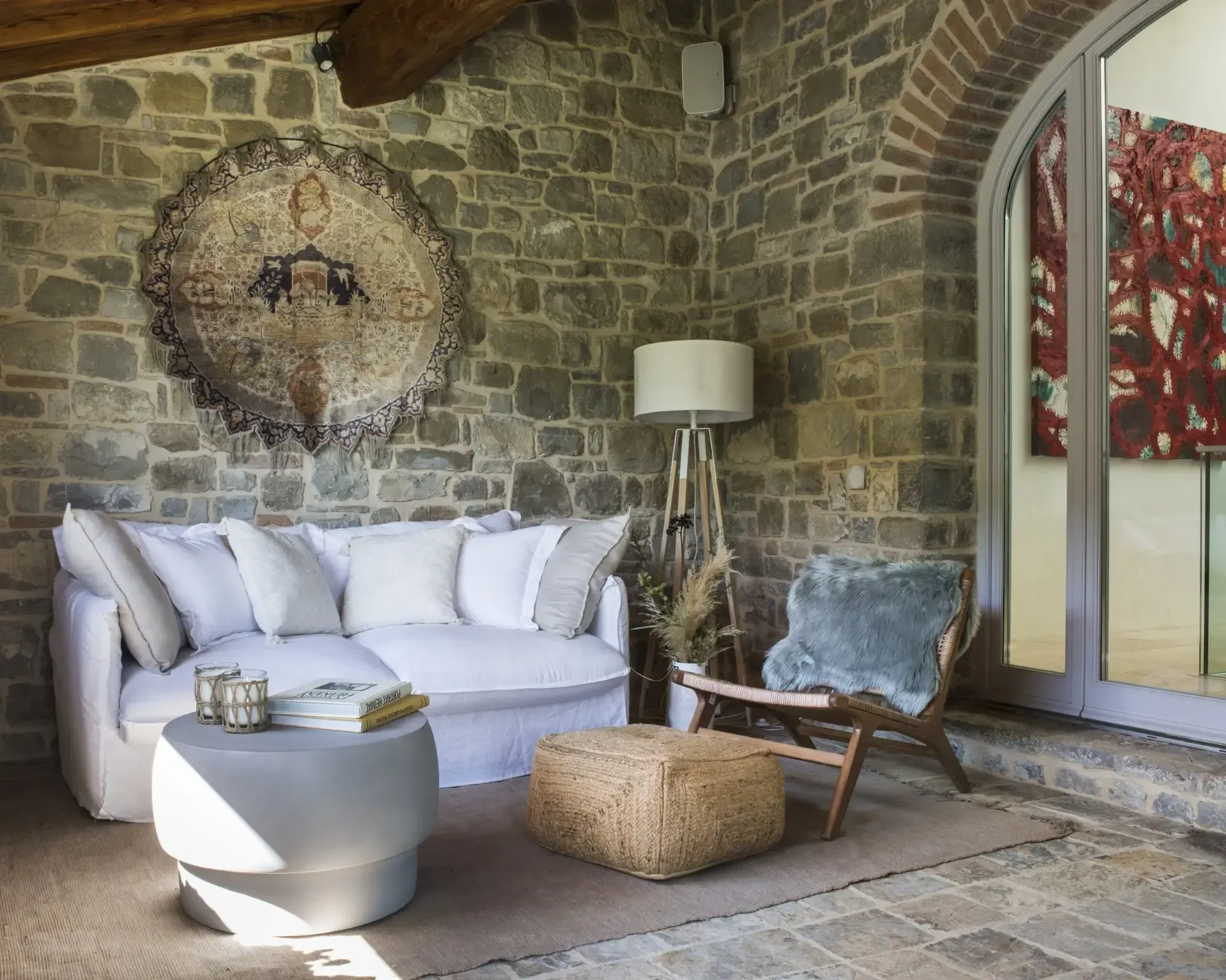 Cozy outdoor seating area with stone wall. White sofa, wooden chair, ottoman, art, and arched doorway.
