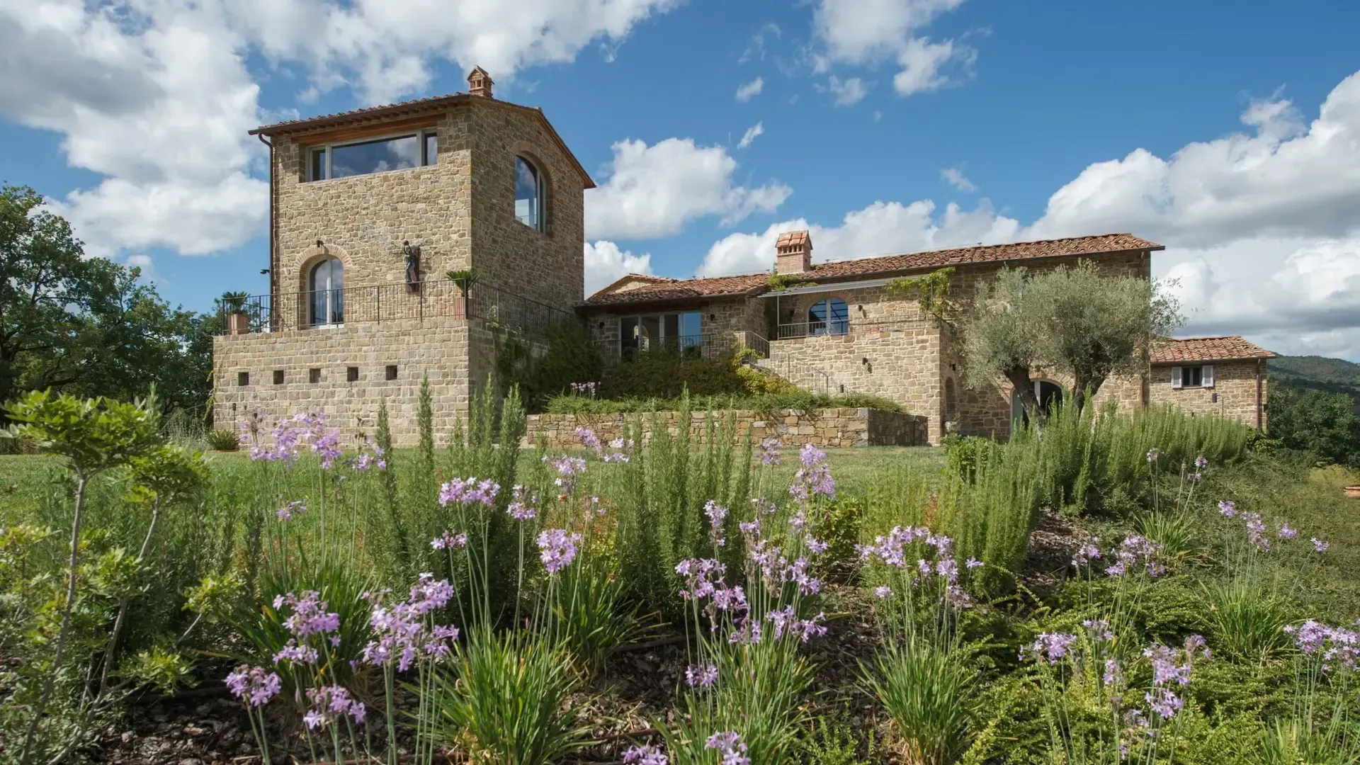 Stone villa with a tall tower and flowering plants under a blue sky.