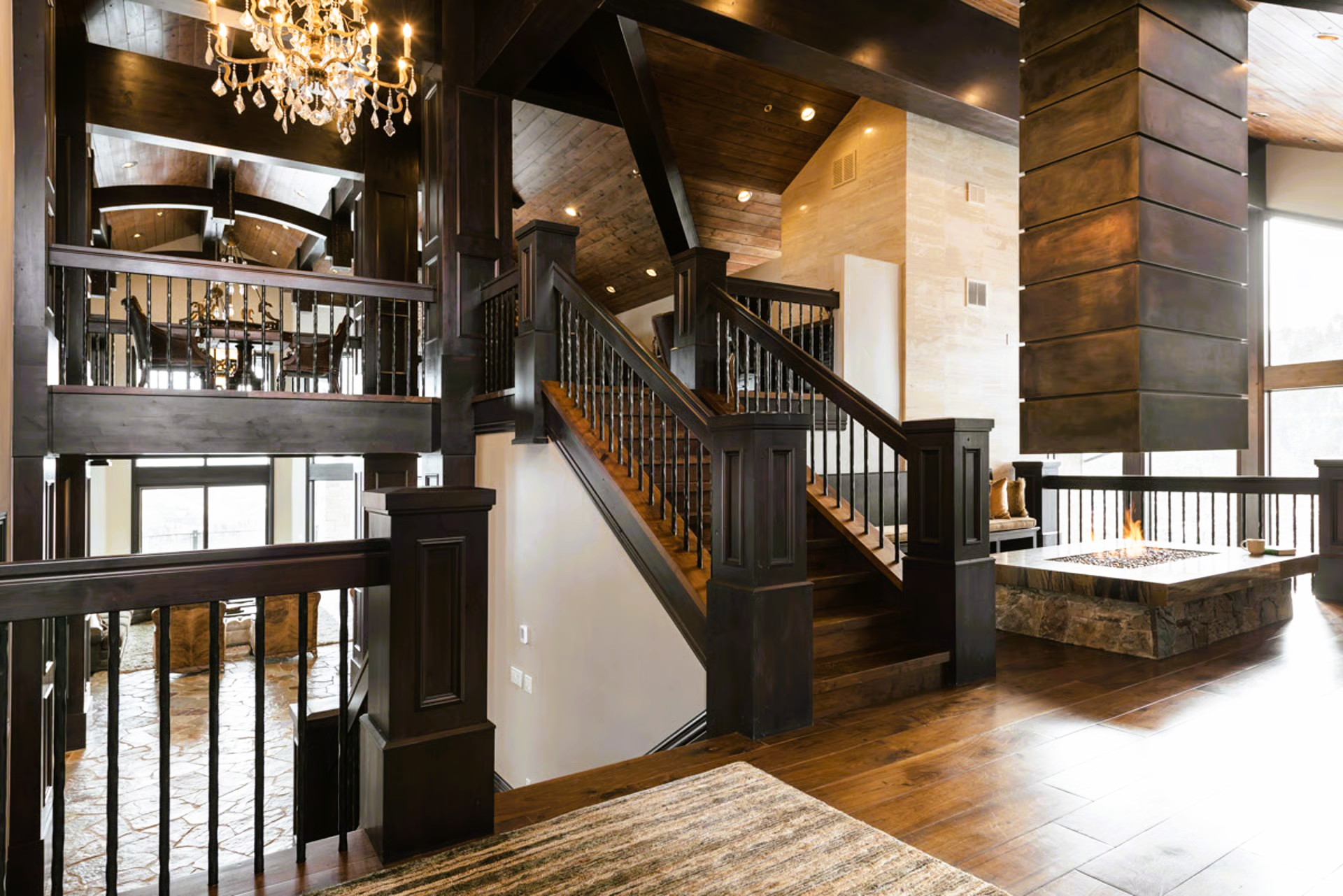 Dark wood staircase in a modern home. Fireplace on the right. Chandelier hanging from ceiling.