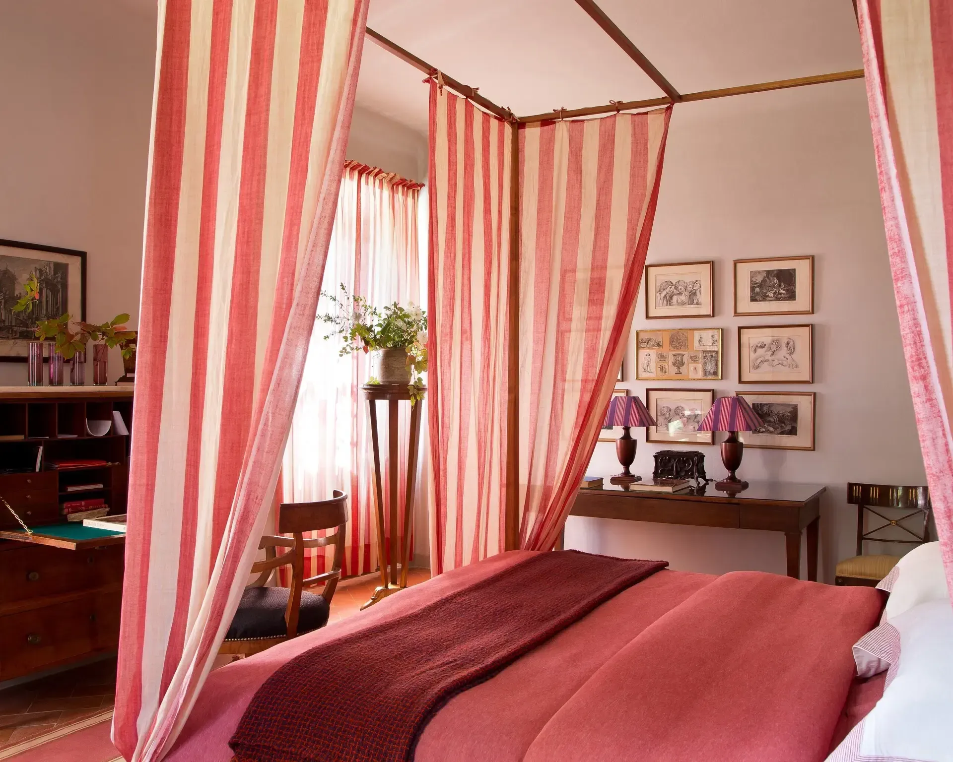 Bedroom with a four-poster bed. Pink and white striped curtains frame the bed. A desk with lamps and artwork on the walls.