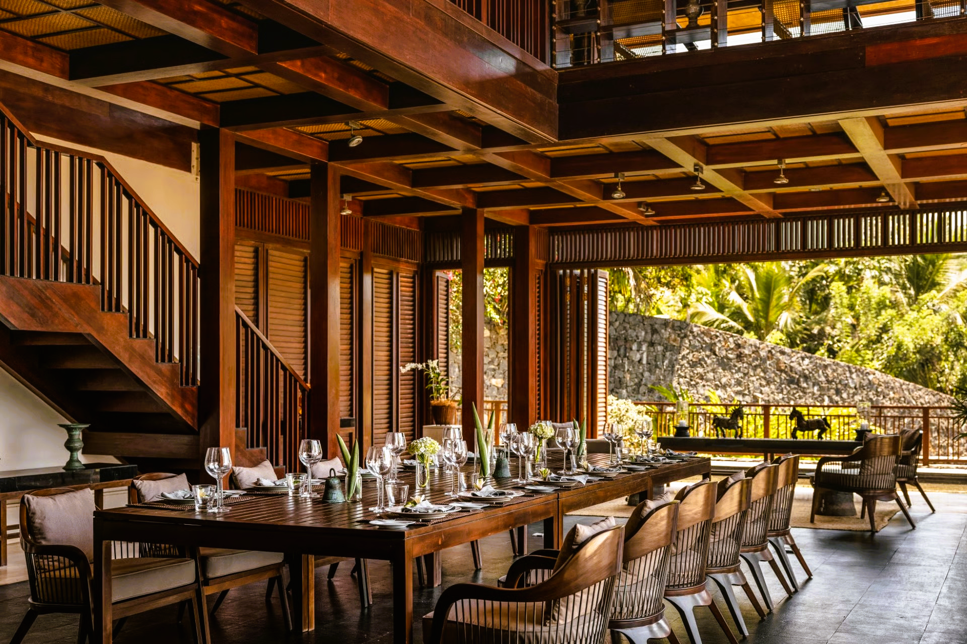 Wooden dining room with long table set for a meal, open to a lush green outdoor view.