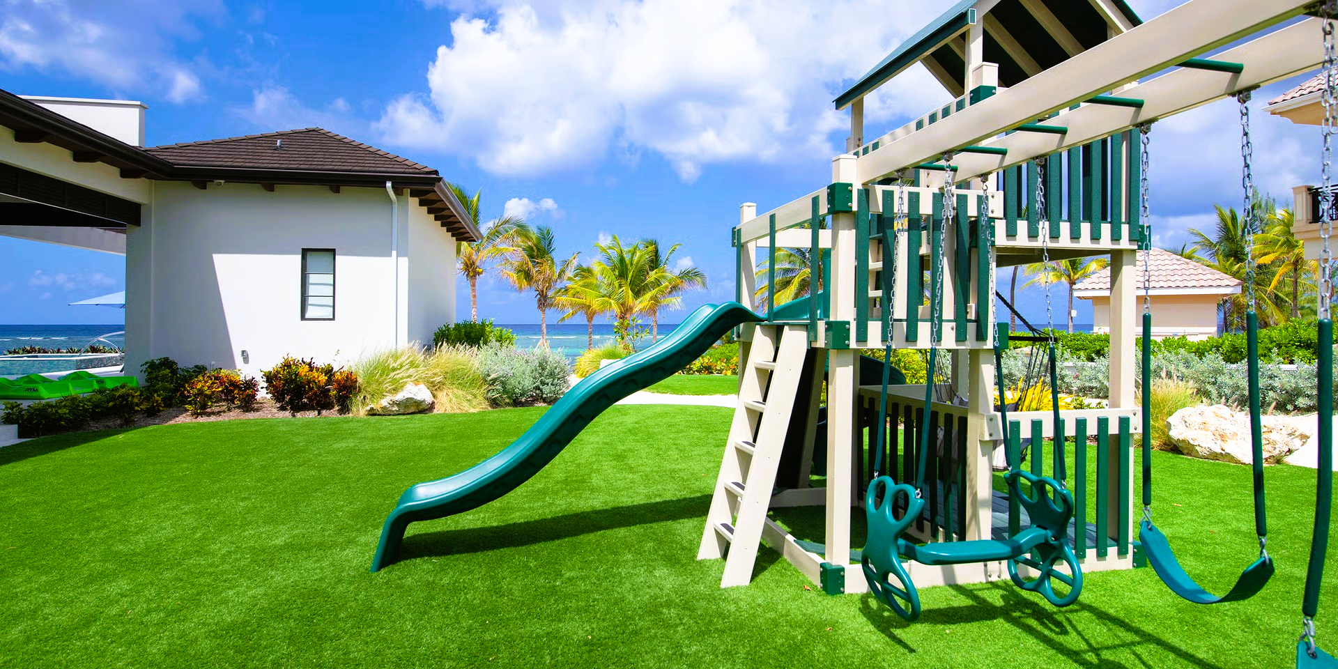 Playground with green slide and swings on a sunny, grassy lawn near a white house.