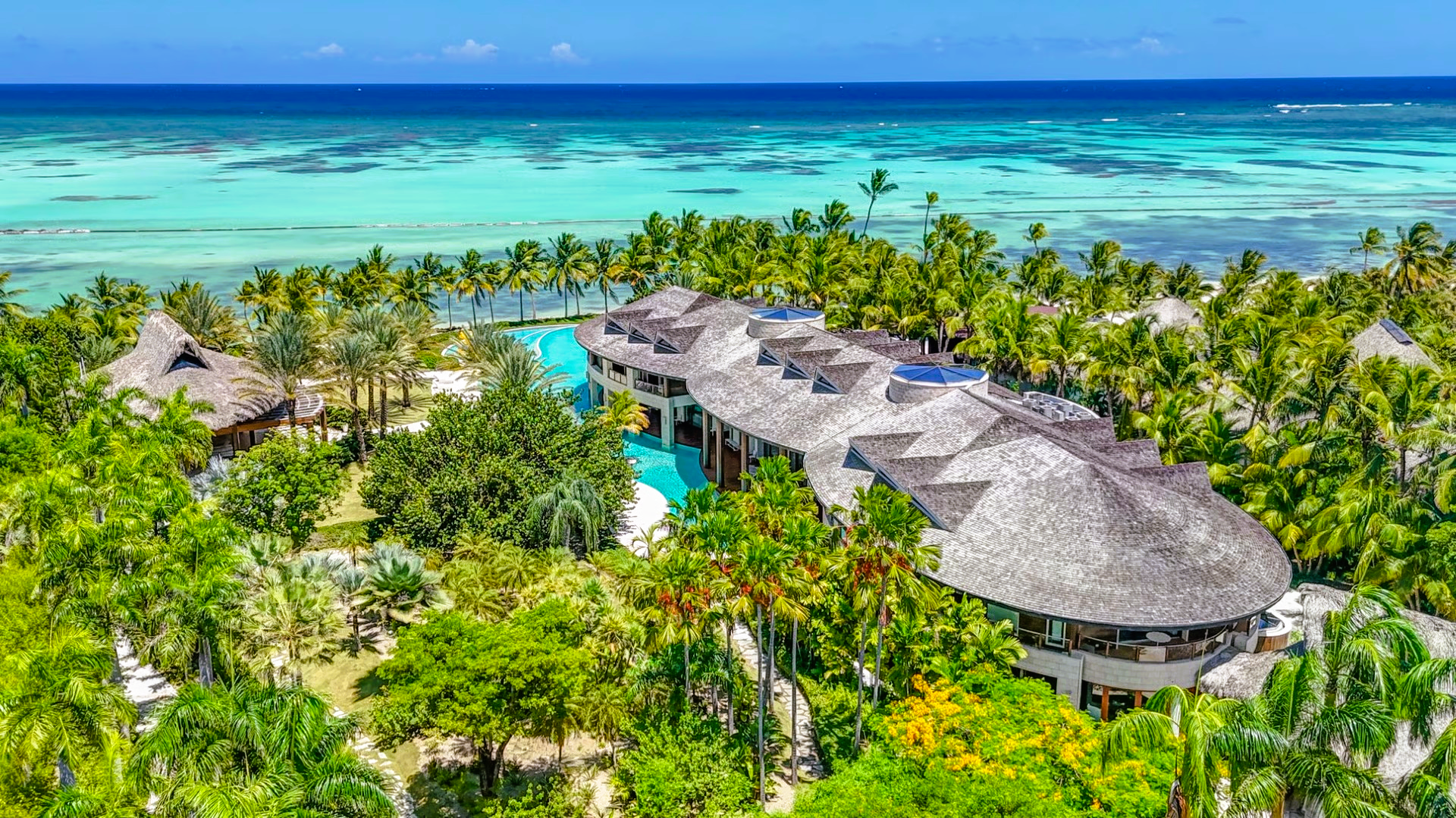 Tropical resort with thatched roofs, pool, lush green trees, and turquoise ocean.