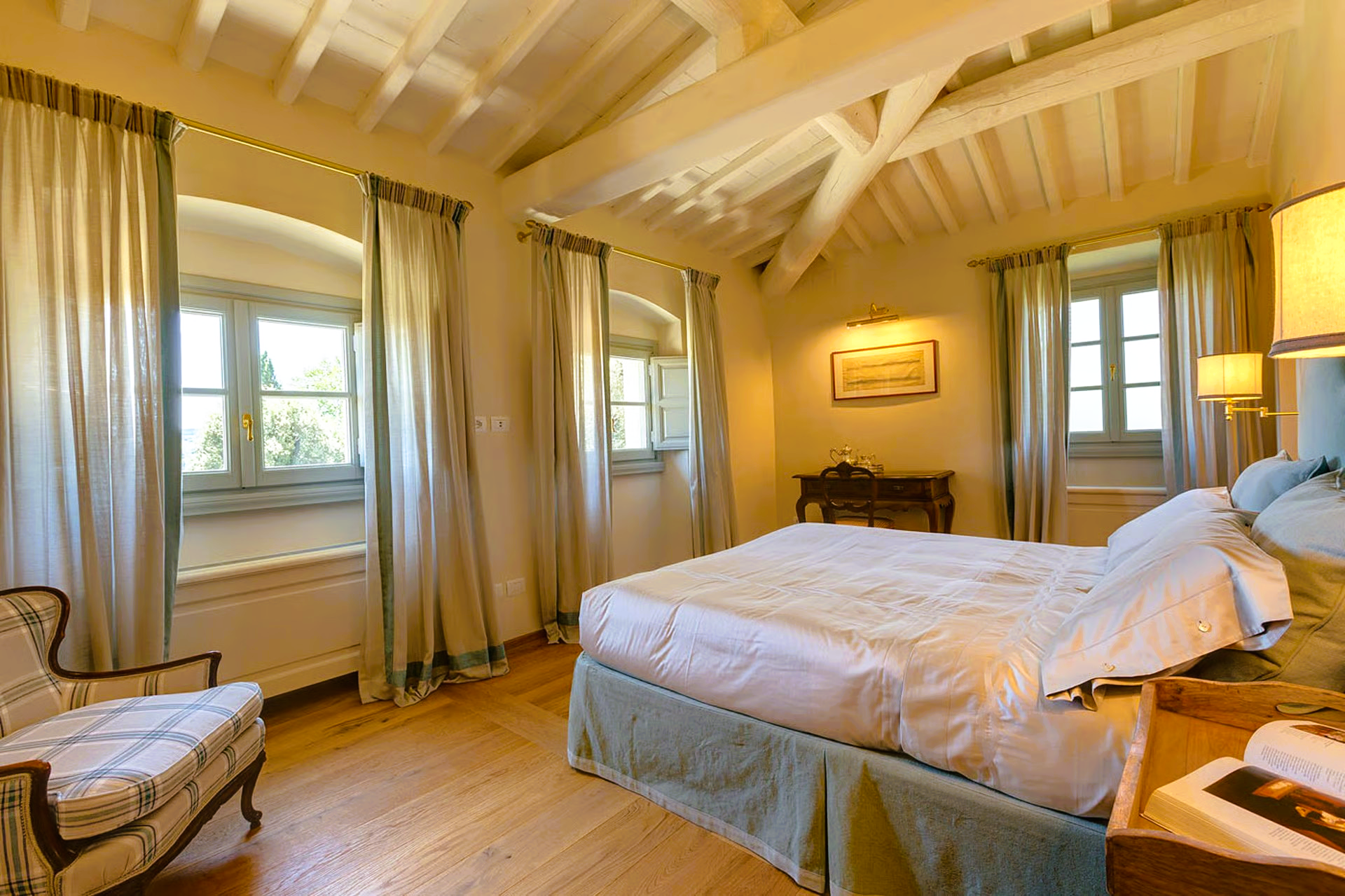Bedroom with white bed, pale curtains, wooden floors, and exposed beams.