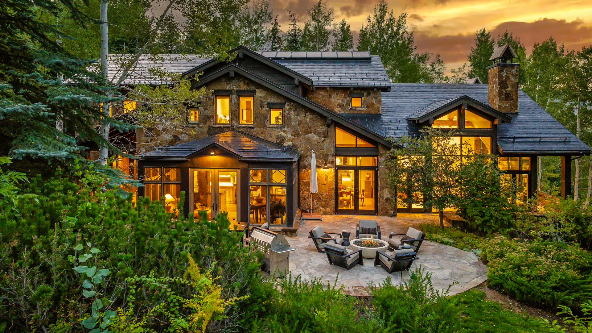 Stone house with lit windows, surrounded by greenery, with a fire pit area.