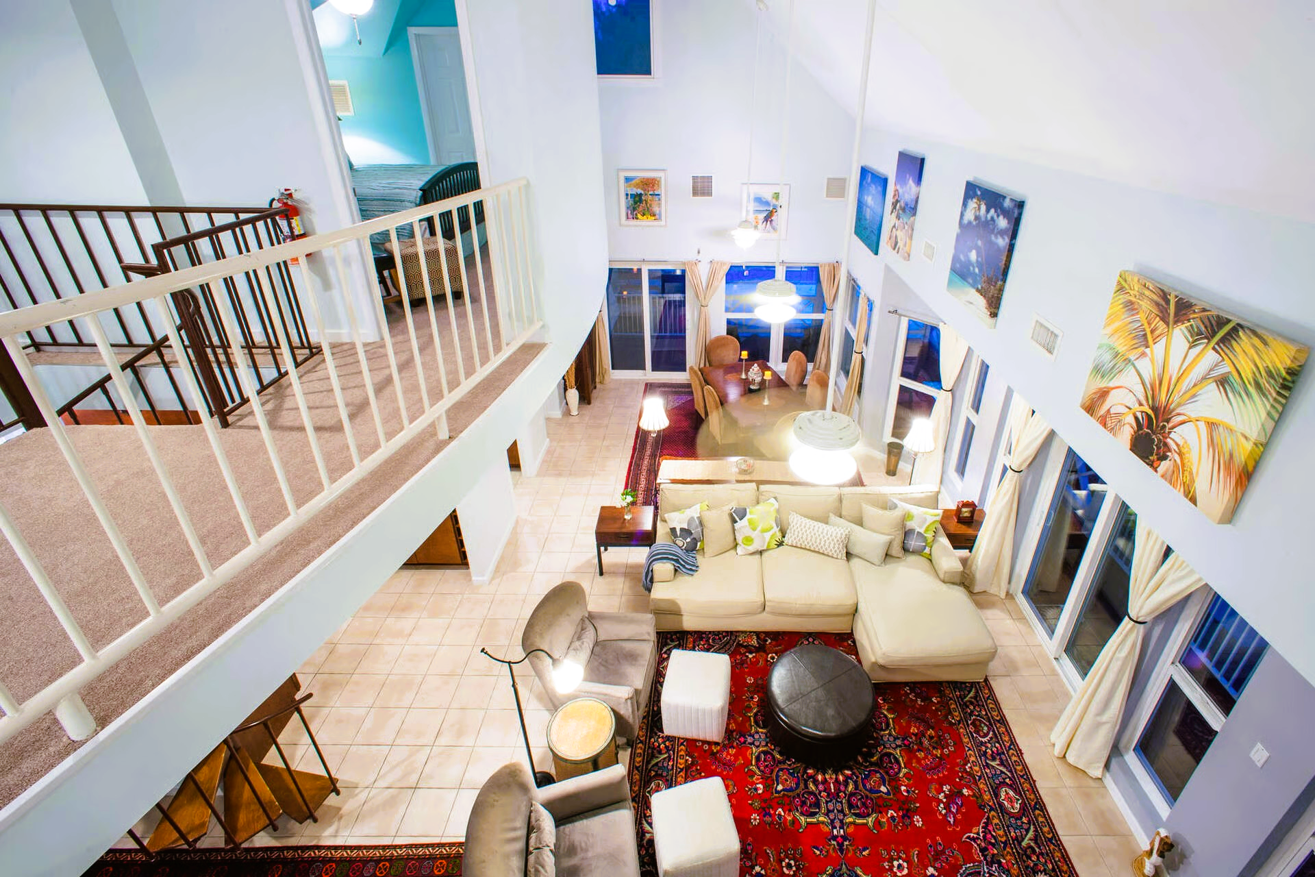 Open, two-story living room. Beige sofa, red rug, paintings, and large windows looking out at the blue sky.