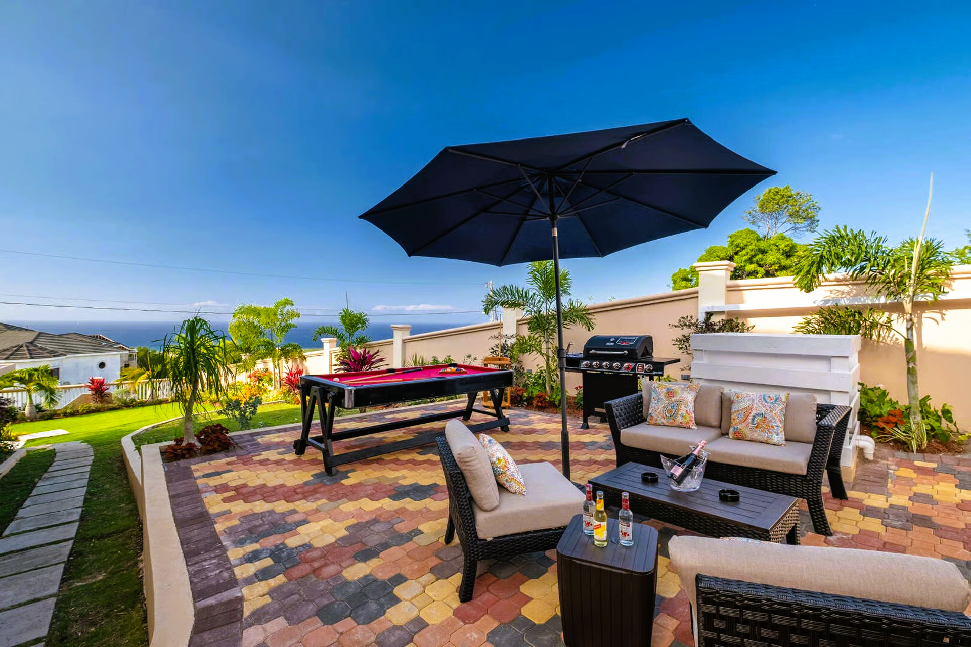 Patio with pool table, seating, grill, and umbrella; ocean view on a sunny day.