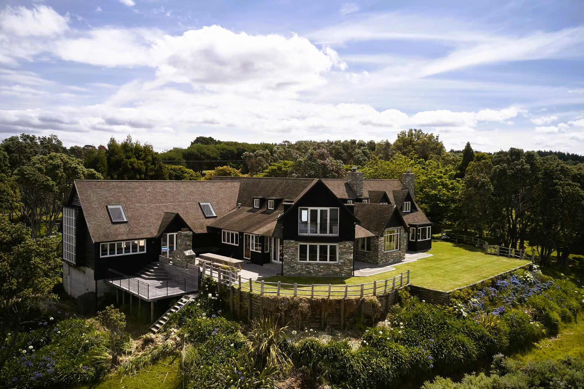 Large house with stone and black exterior, set on a hill with lush greenery.