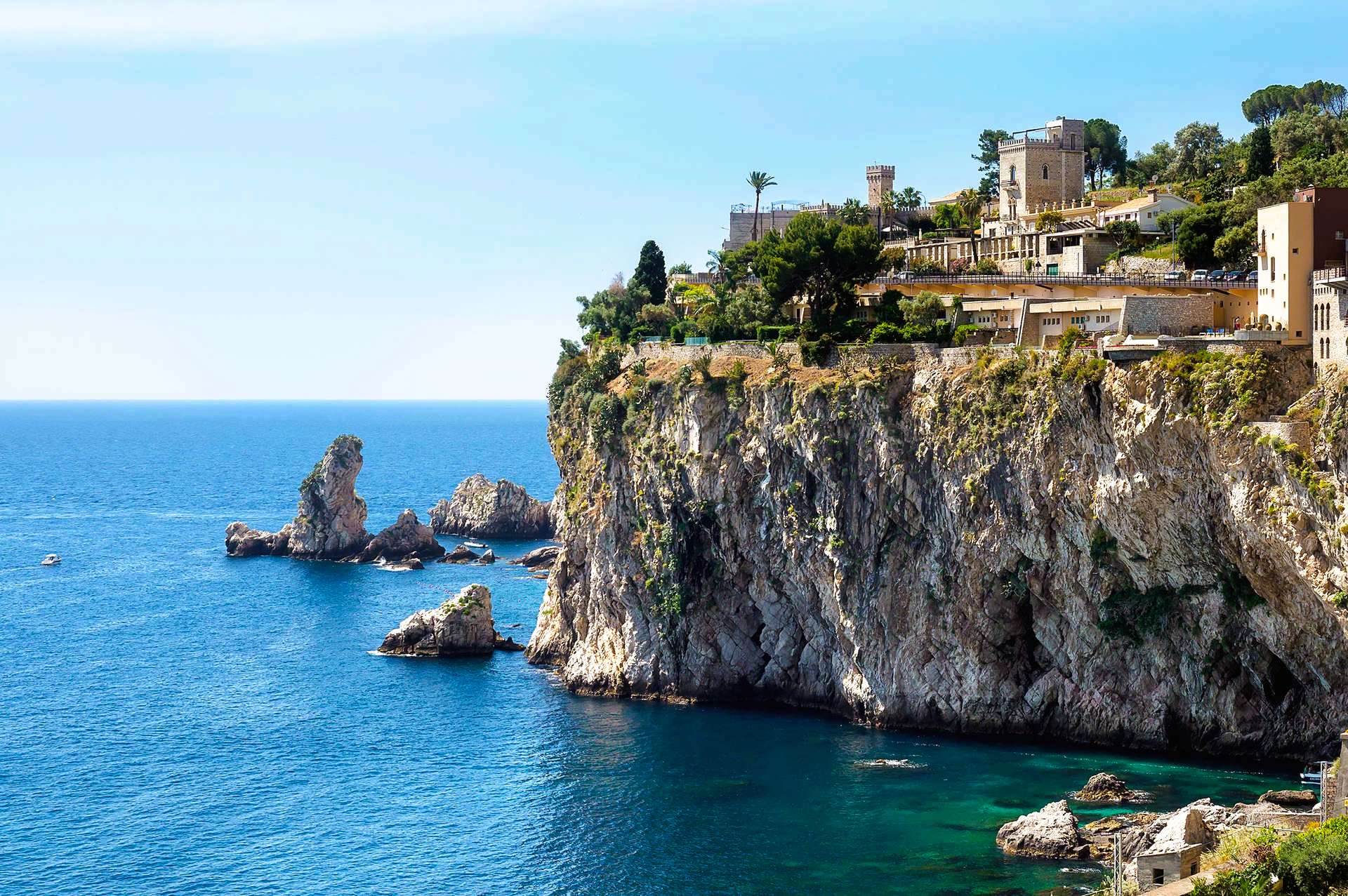 Cliffs with buildings overlook a blue sea. Small islands and boats visible.