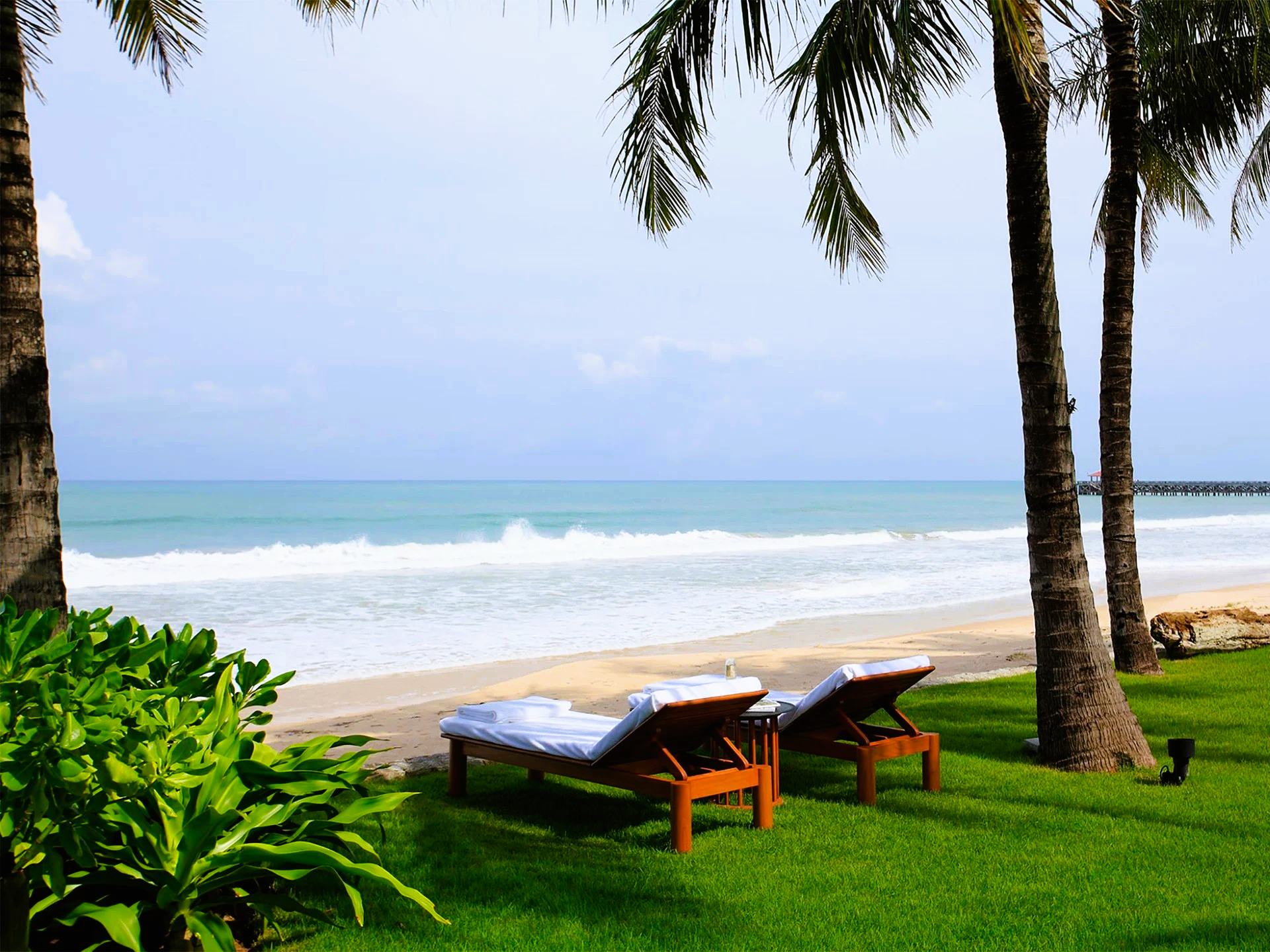 Two lounge chairs on a green lawn overlooking a sandy beach and ocean under a blue sky.
