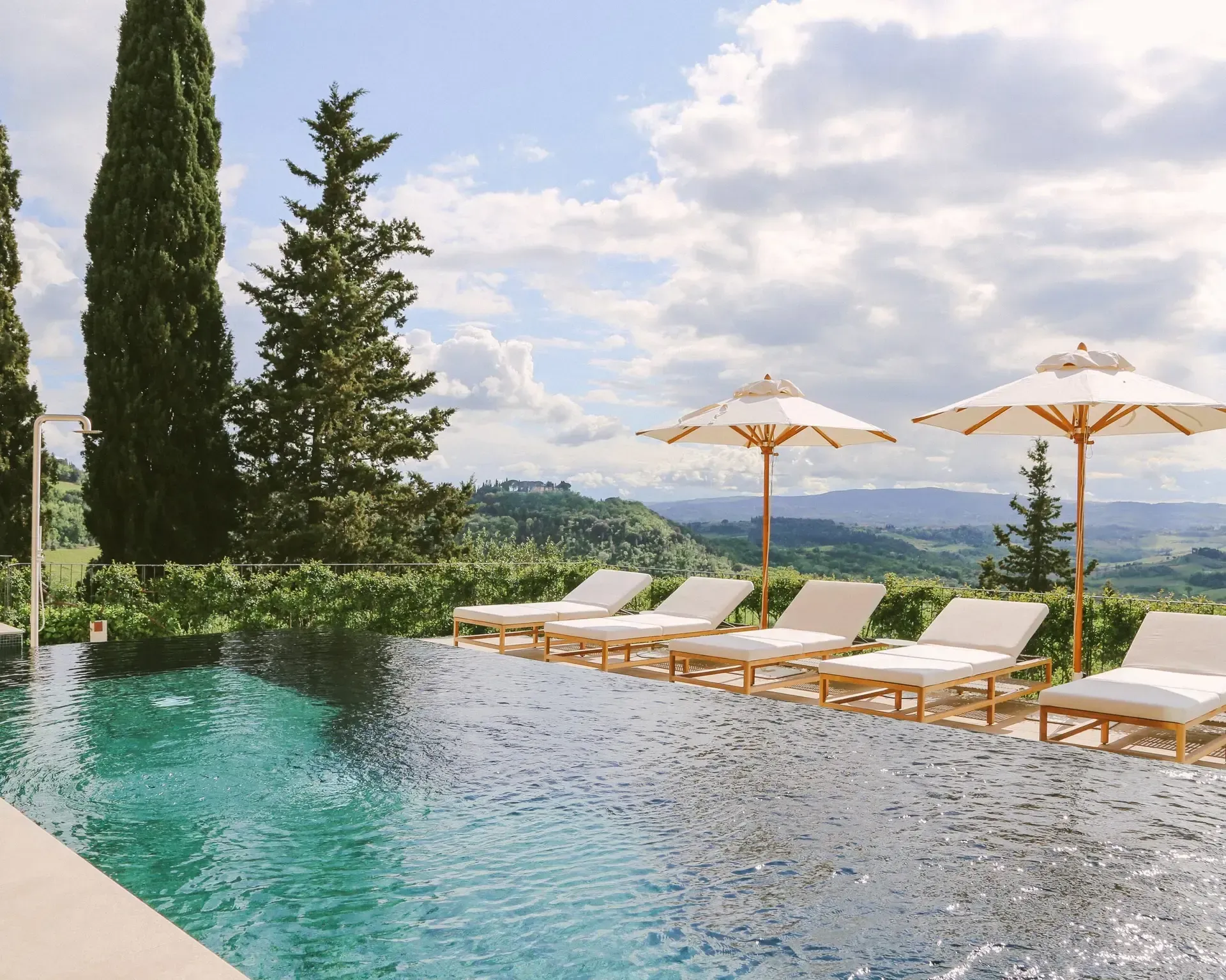 Infinity pool with lounge chairs, umbrellas, overlooking Tuscan hills on a cloudy day.