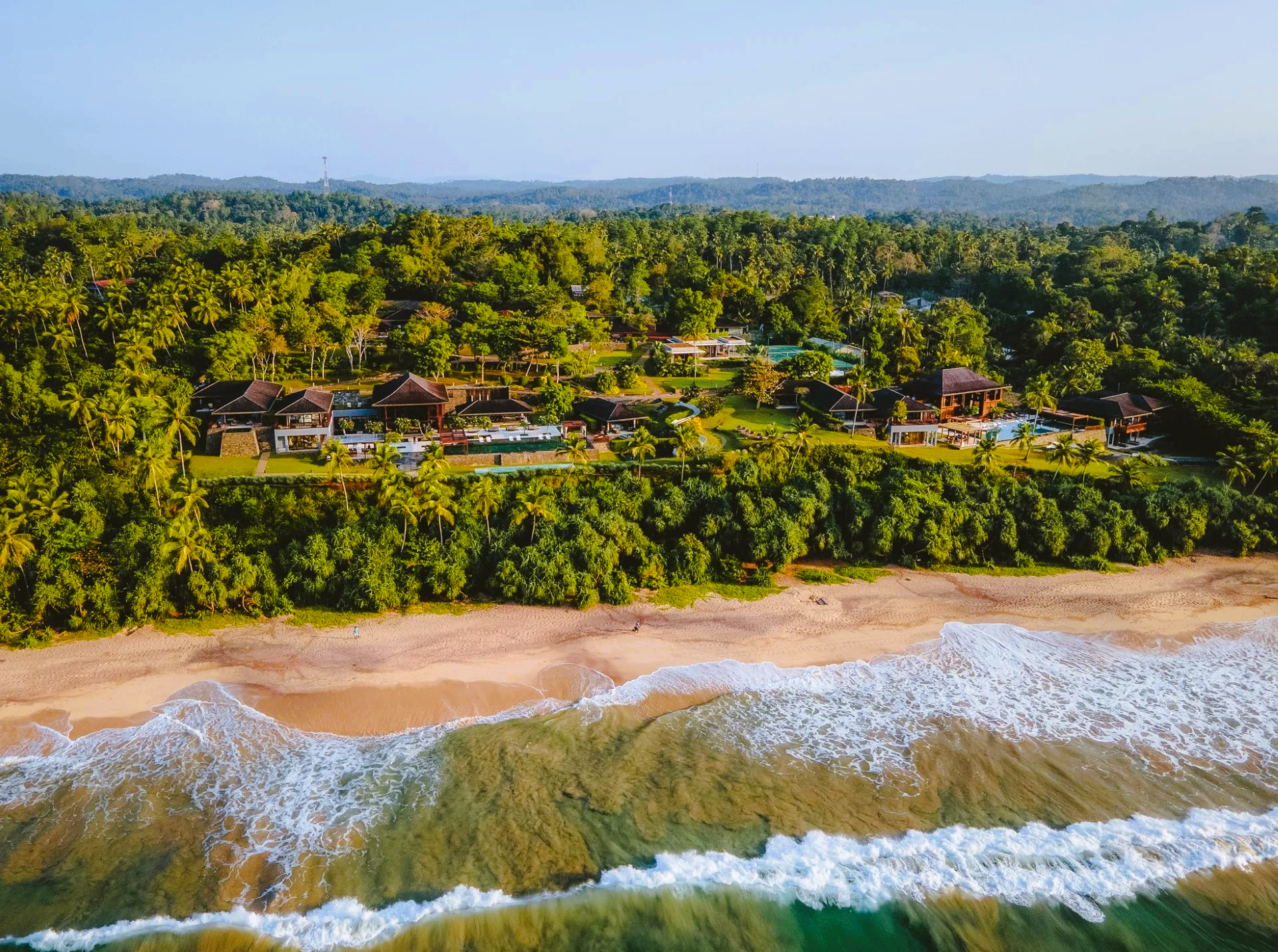 Aerial view of a beach with waves, lush green forest, and buildings nestled along the coast.