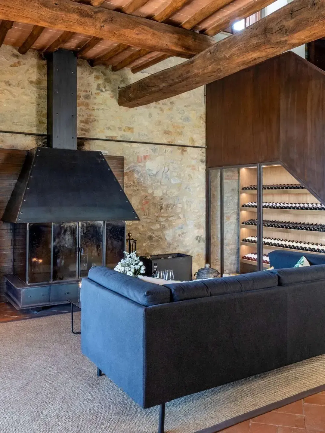 Cozy living room with fireplace, dark blue sofa, and exposed beams.  Wine rack on the right.  Brick wall and stone flooring.