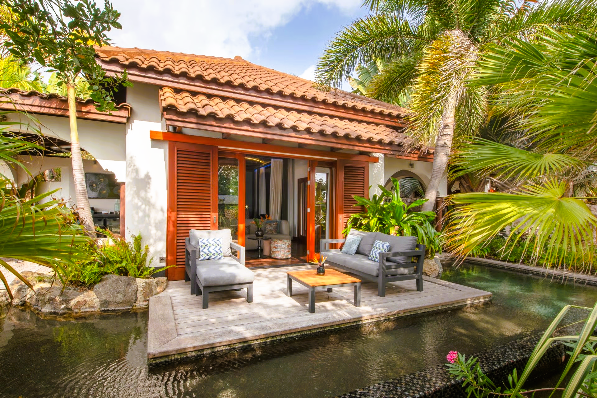 Small white building with terracotta roof and wood trim, opens onto a patio over a pond.