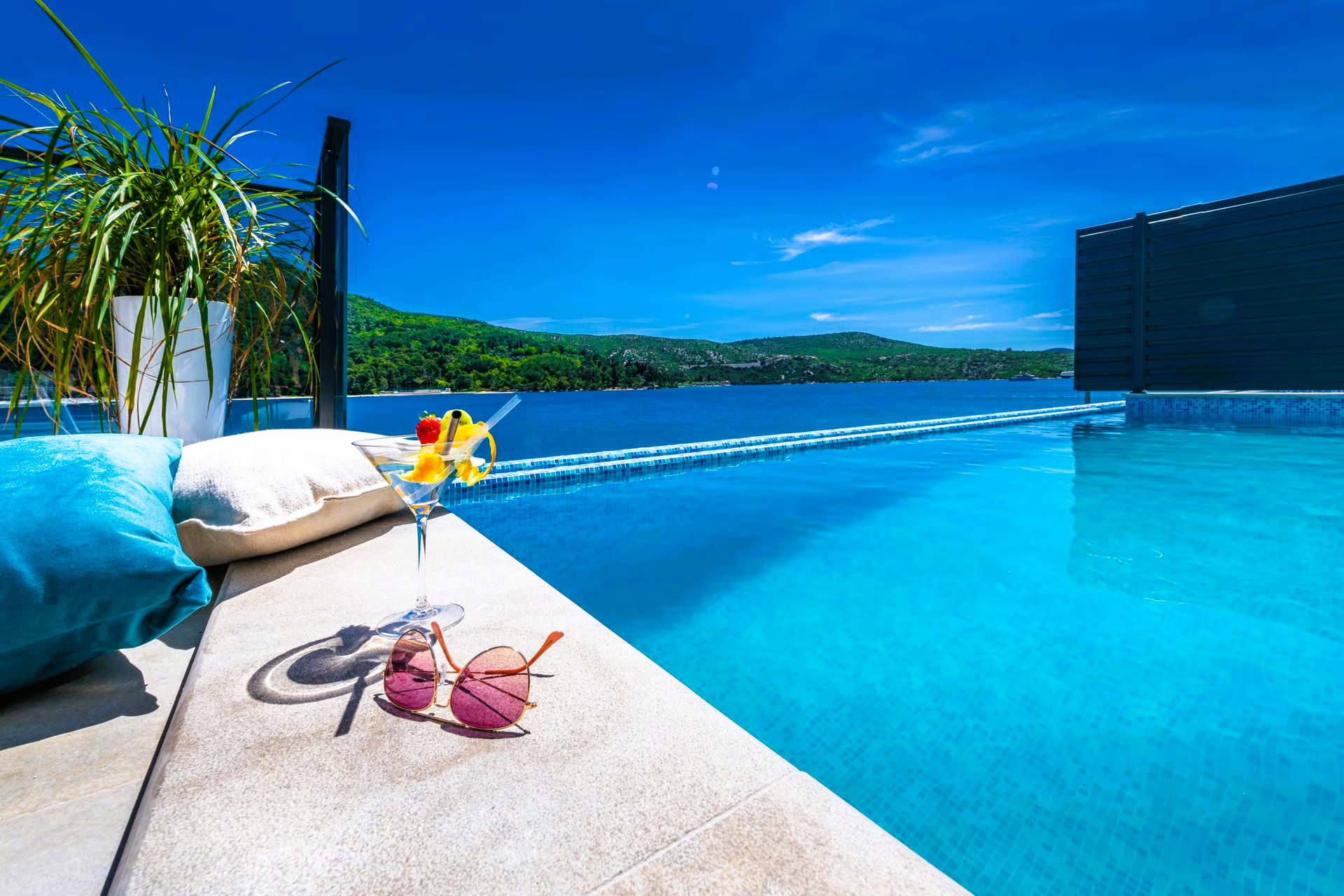 Poolside scene with a cocktail and sunglasses, overlooking a blue ocean and mountains.