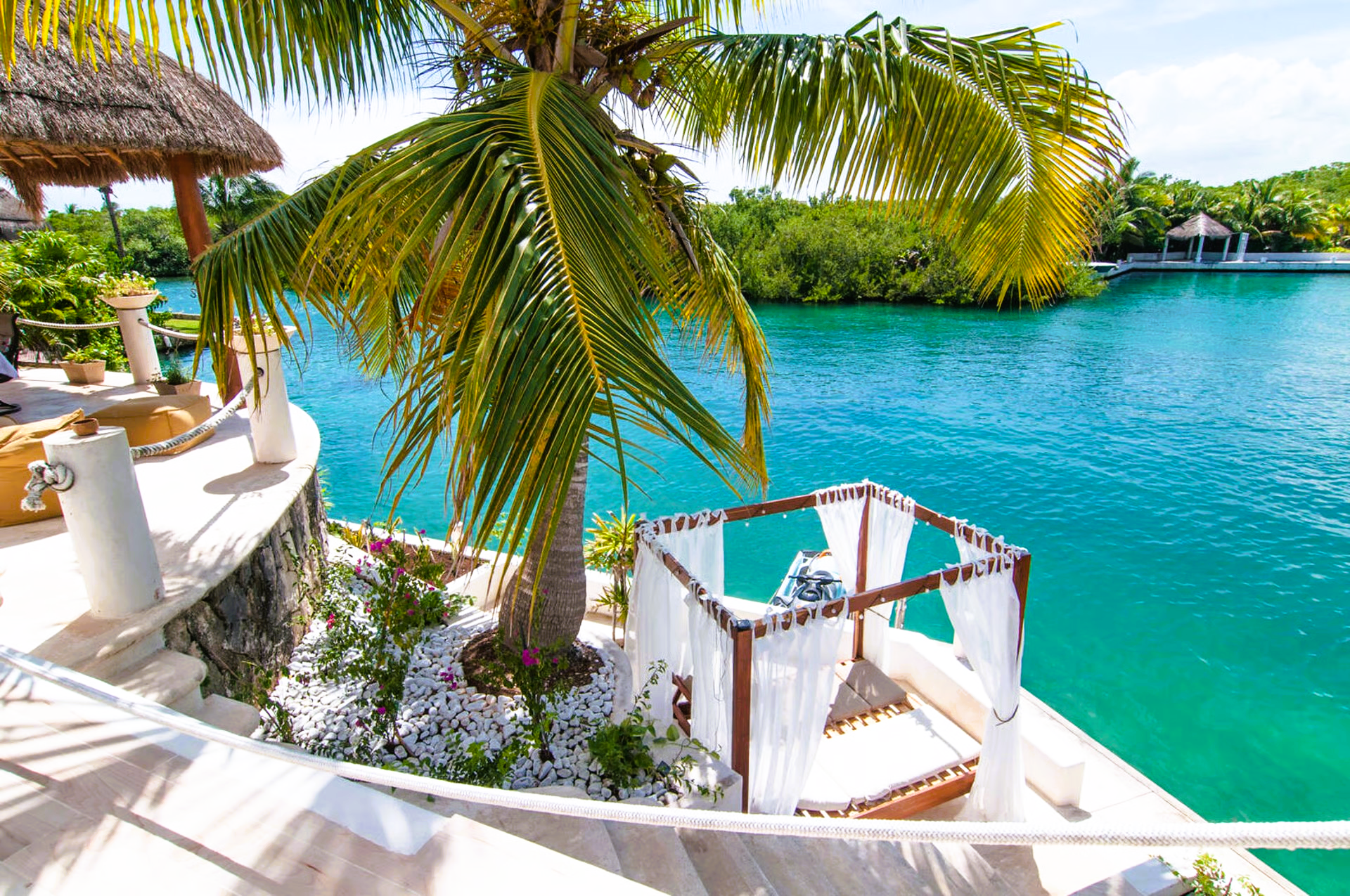 Palm tree over a white gazebo with flowing curtains near turquoise water and lush greenery in a tropical setting.