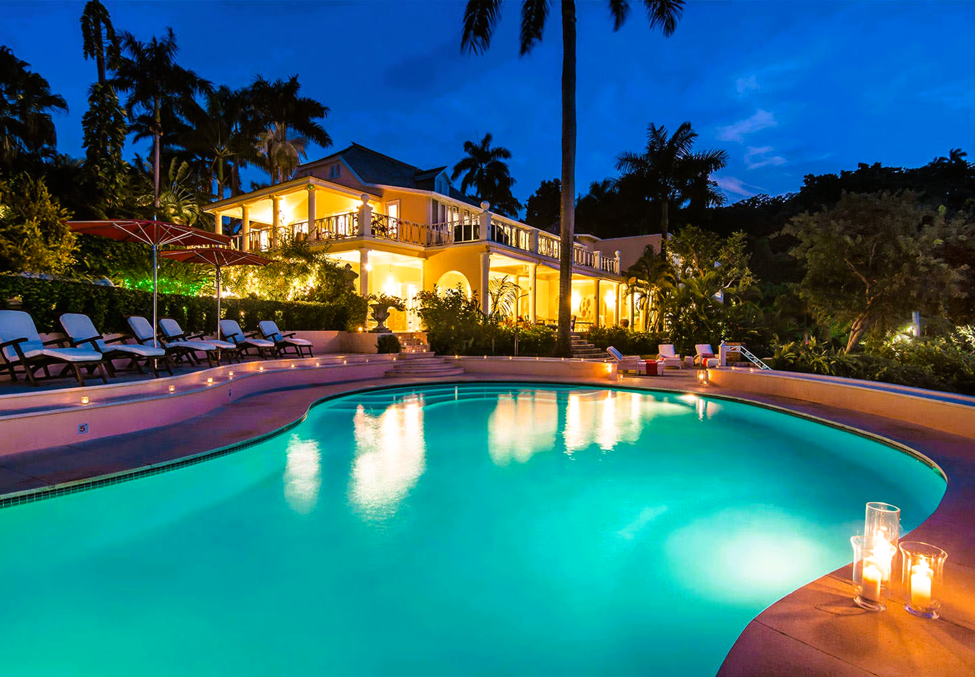 Nighttime view of a pool illuminated by lights, next to a luxurious two-story building with palm trees.