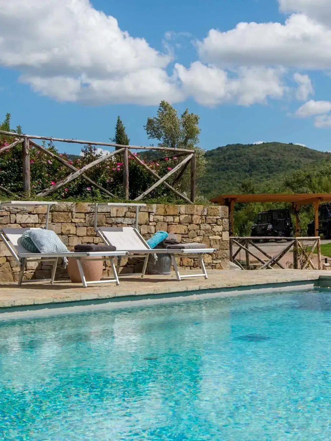 Swimming pool with lounge chairs, stone wall, wooden fence, and green hills under a blue sky.