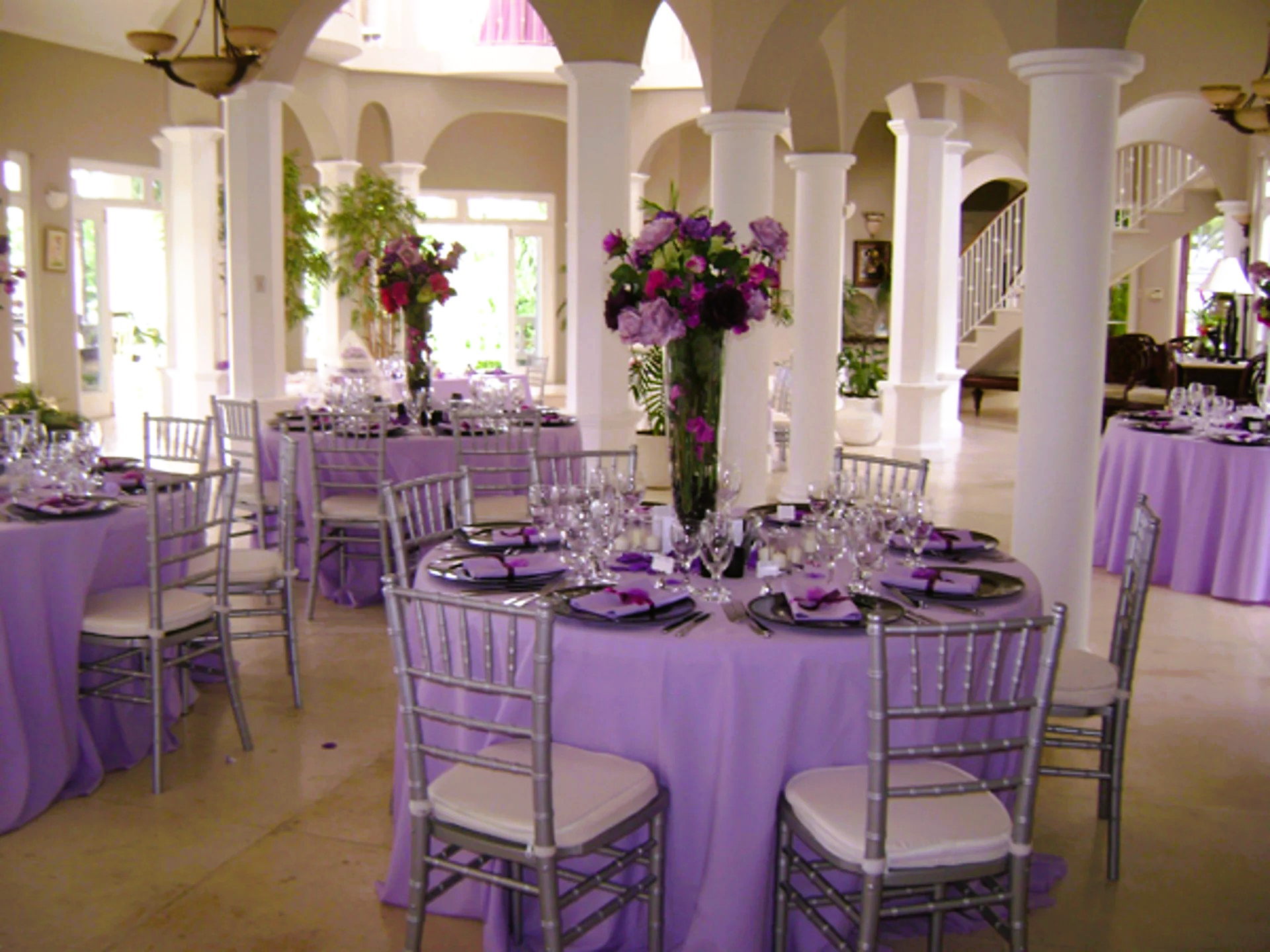 Wedding reception with lavender tablecloths, silver chairs, and floral centerpieces in a bright, open space.