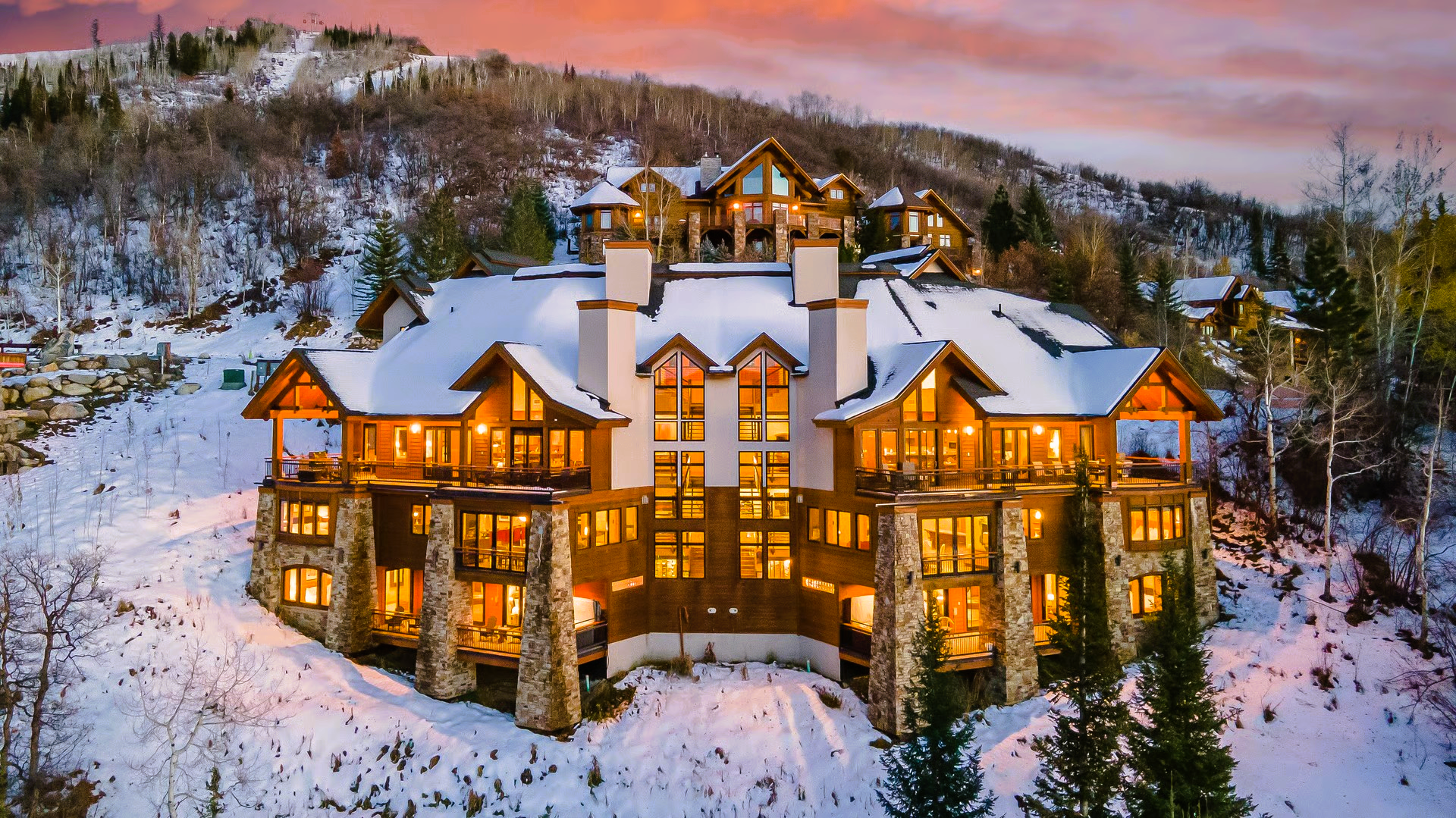 Luxury snow-covered cabin with lit windows, on a mountain at sunset.