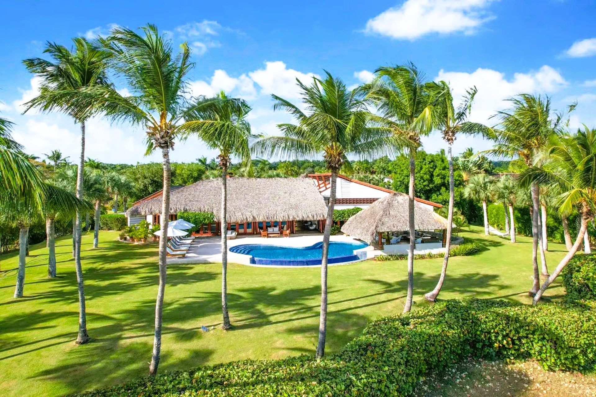 Tropical villa with pool, lush green lawn, palm trees, and blue sky.