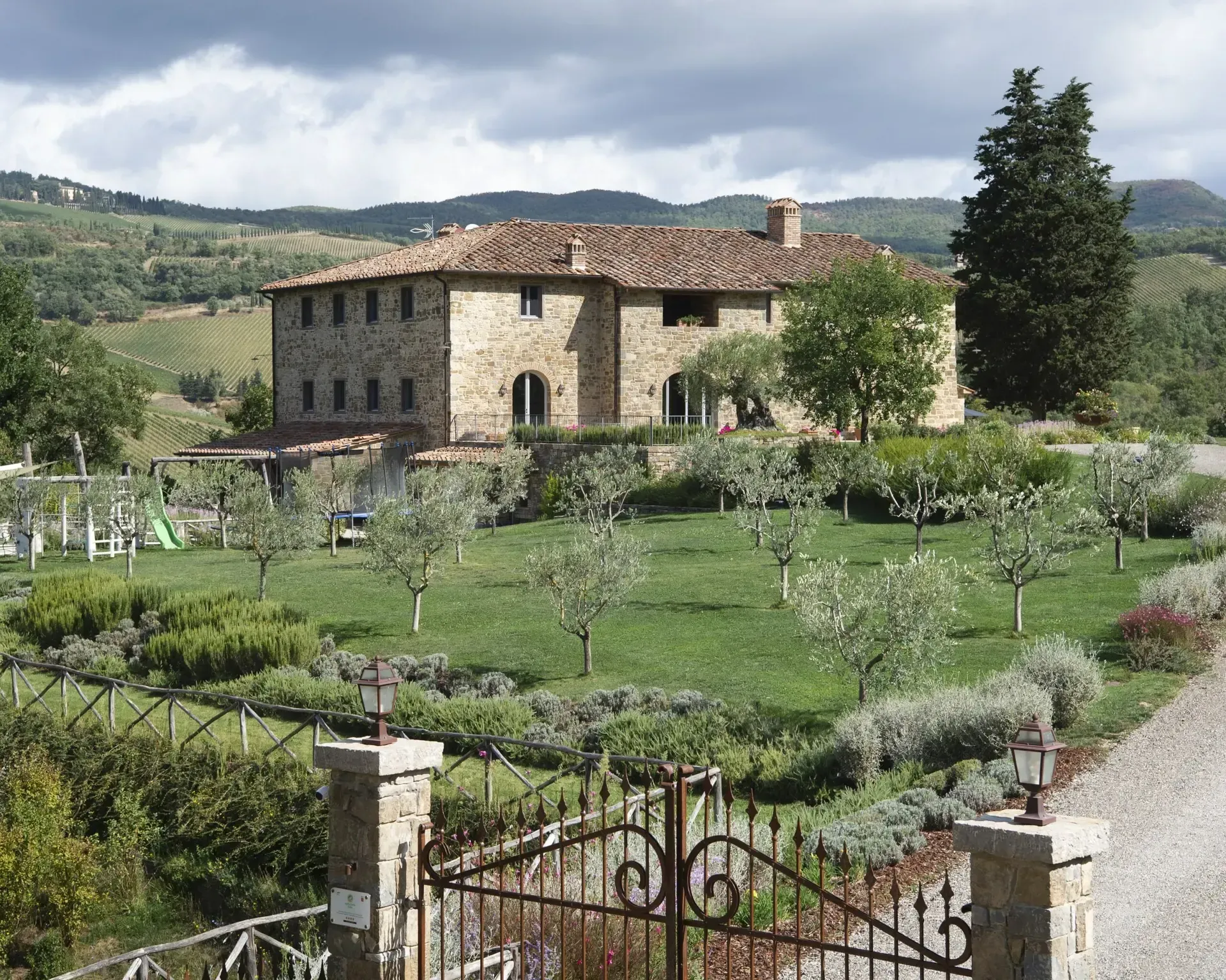 Stone house with tile roof and arched windows, set in a Tuscan landscape with a manicured garden and rolling hills.