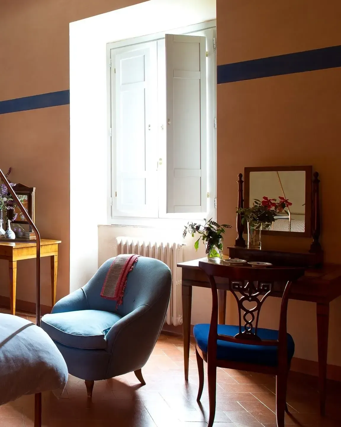 A sunlit room with blue armchair, wooden table and chair, a window and flowers.