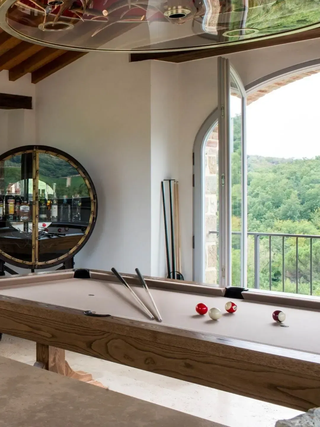 Pool table in a room with a large window overlooking a green landscape. Cue sticks, and a round cabinet visible.