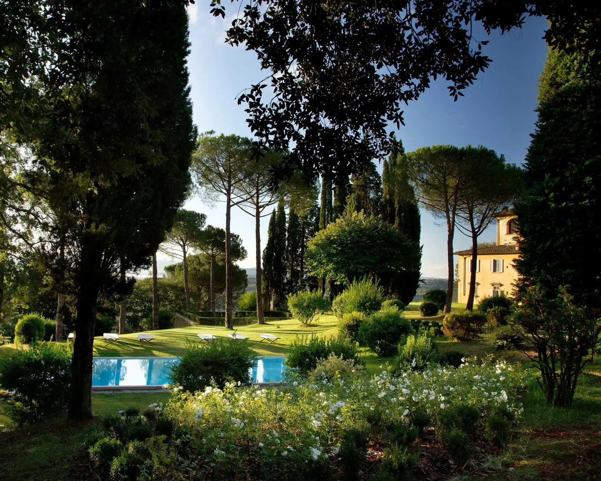 A garden with a pool, trees, and a yellow building in the distance.