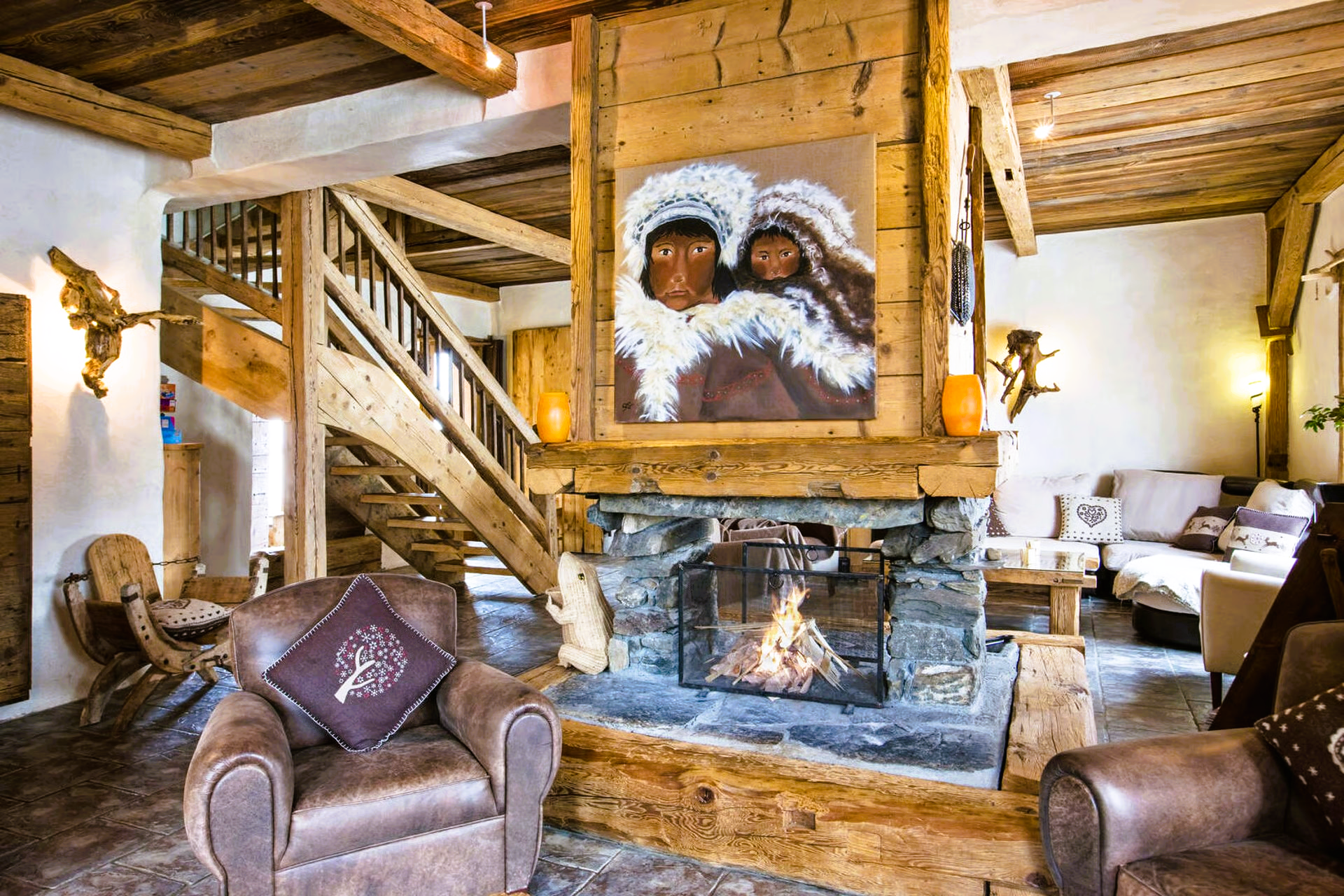 Cozy chalet living room with stone fireplace, wooden beams, leather chairs, and a painting above.
