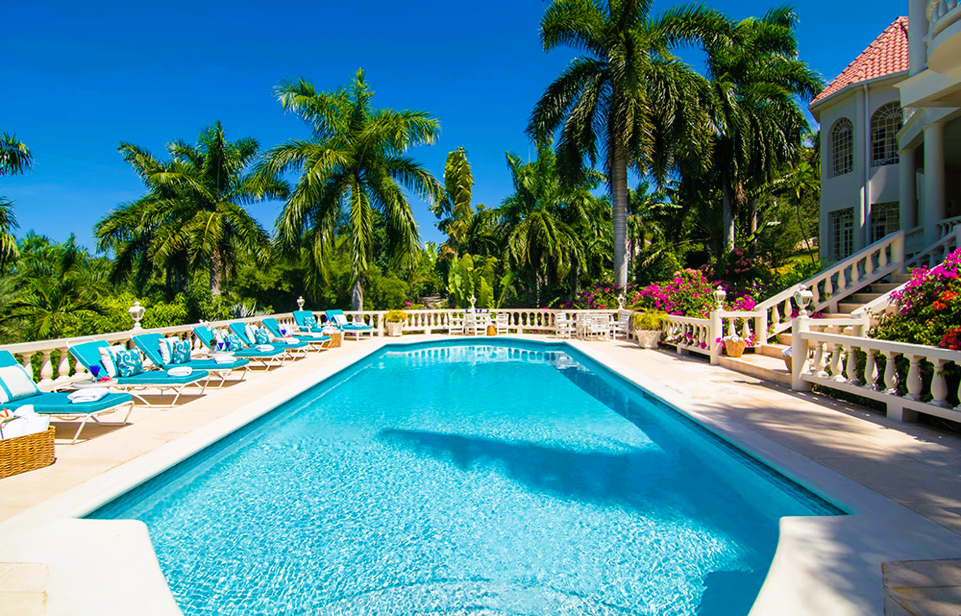 Swimming pool surrounded by lounge chairs, palm trees, and a mansion on a sunny day.