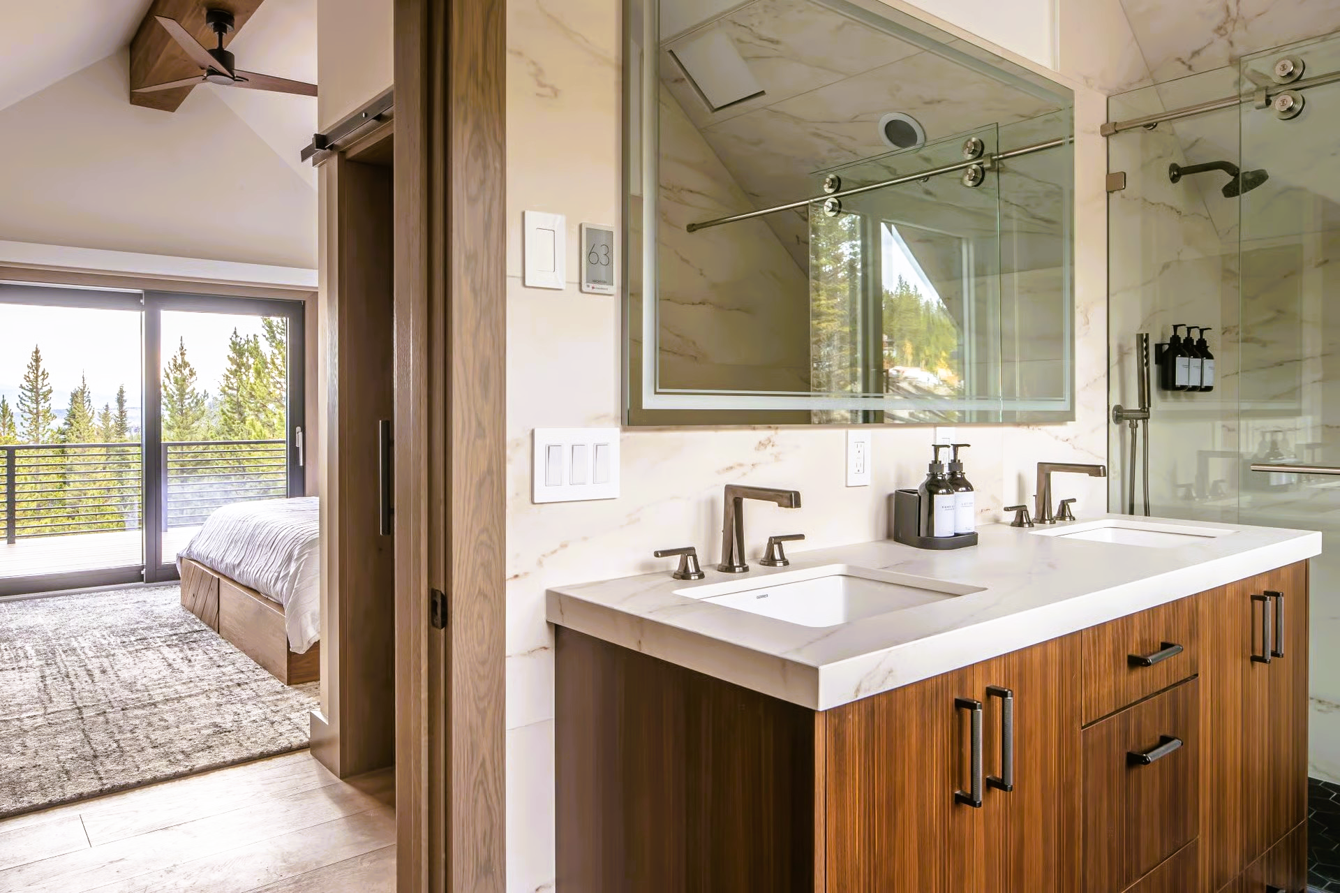 Bathroom with double vanity, marble tile, glass shower, and a bedroom with forest views visible.