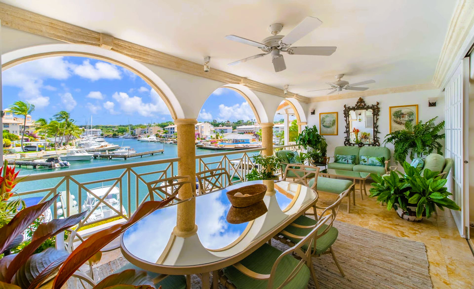 Balcony overlooking a marina with dining table, green chairs, and potted plants; blue sky and water.