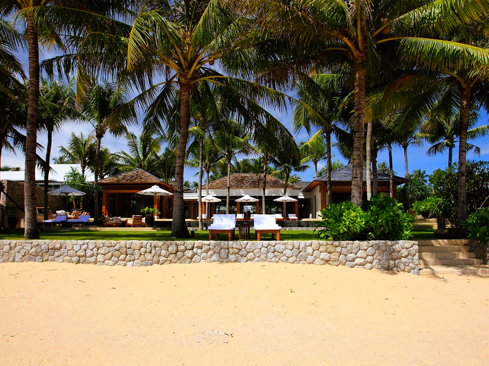 Beachfront resort with palm trees, lounge chairs, and cabanas on a sunny day.