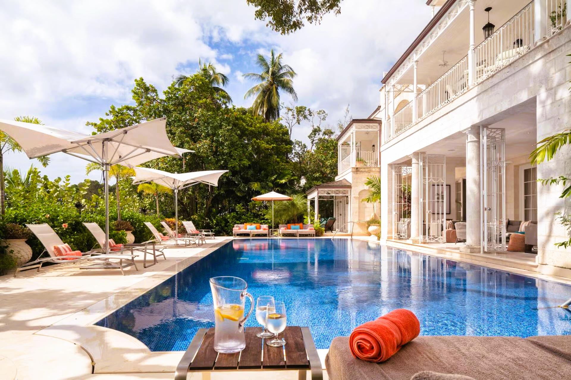 Poolside view of a white villa with a blue-tiled pool, sun loungers, and a table with drinks.