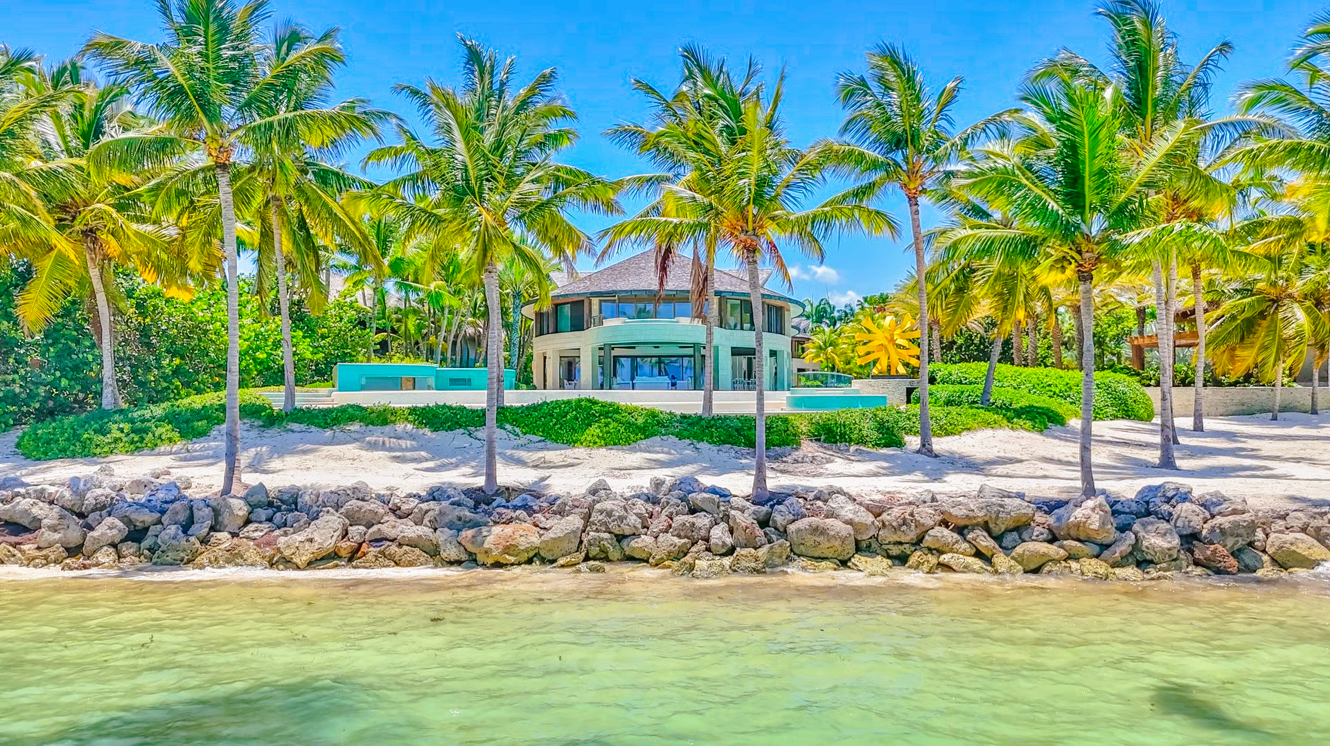 Beachfront mansion with palm trees, turquoise water, and blue sky.