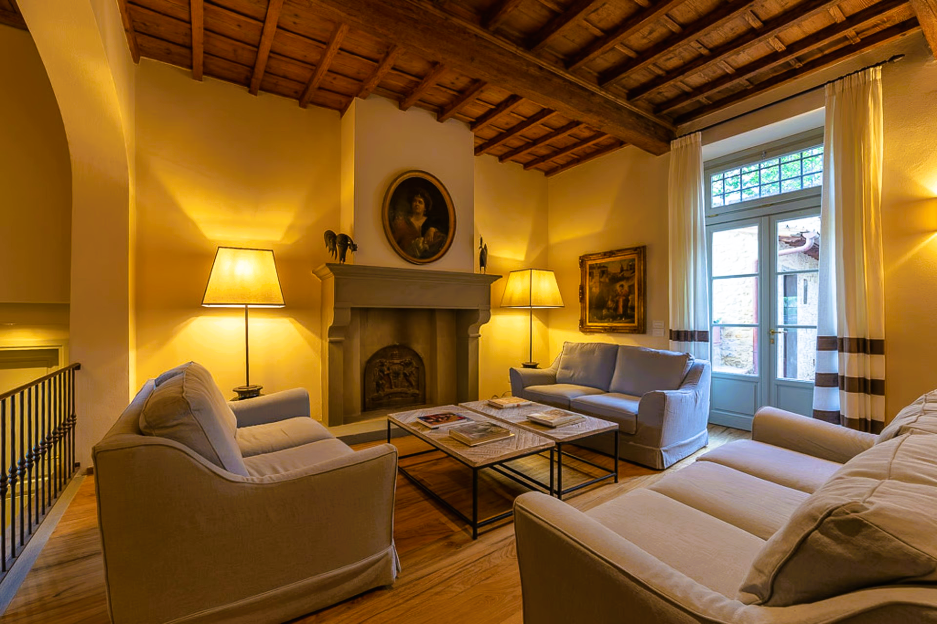 Cozy living room with sofas, fireplace, and lamps. Yellow walls, wooden ceiling, and a view out a window.