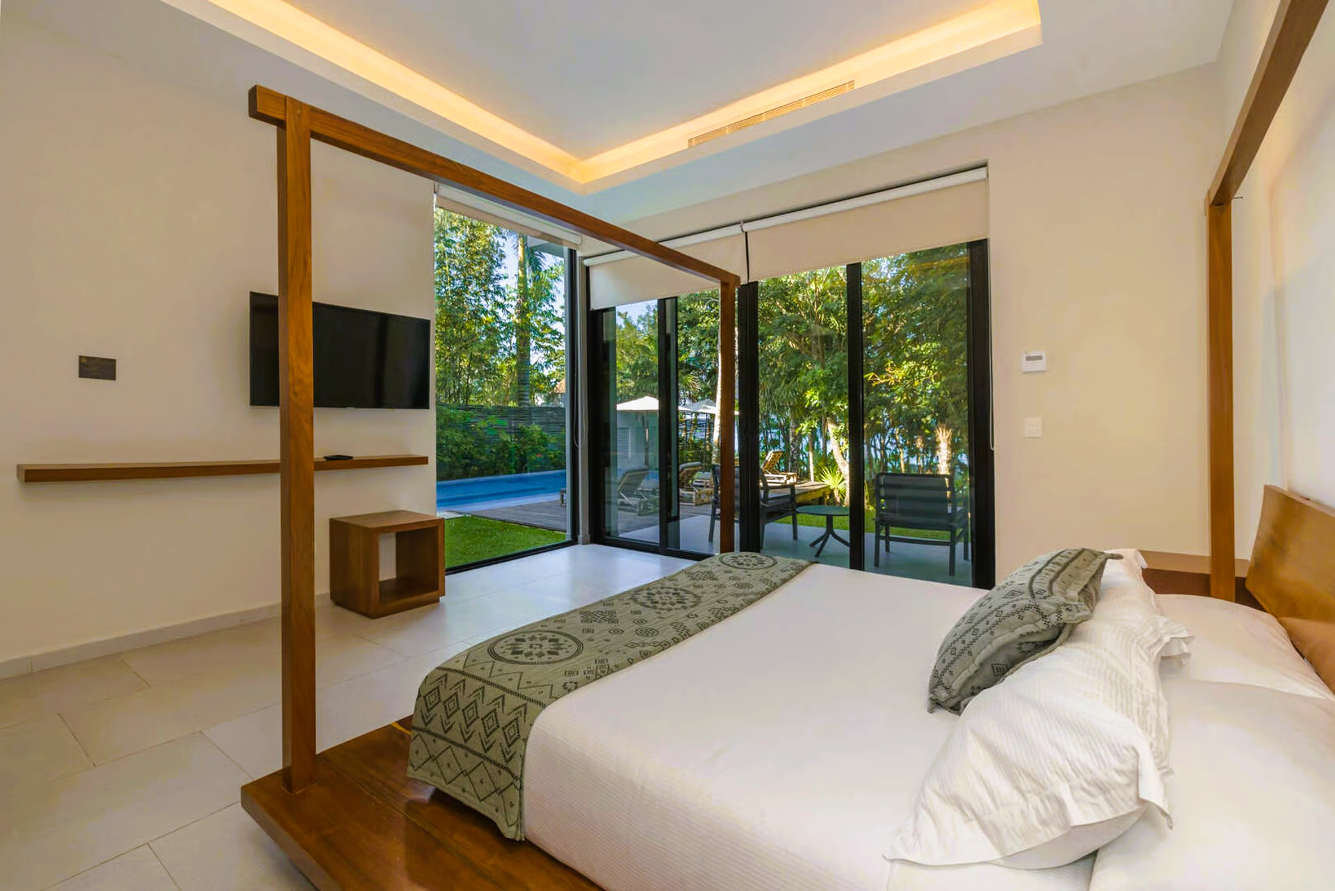 Bedroom with wooden bed frame, glass doors to yard, and a pool visible through the trees.