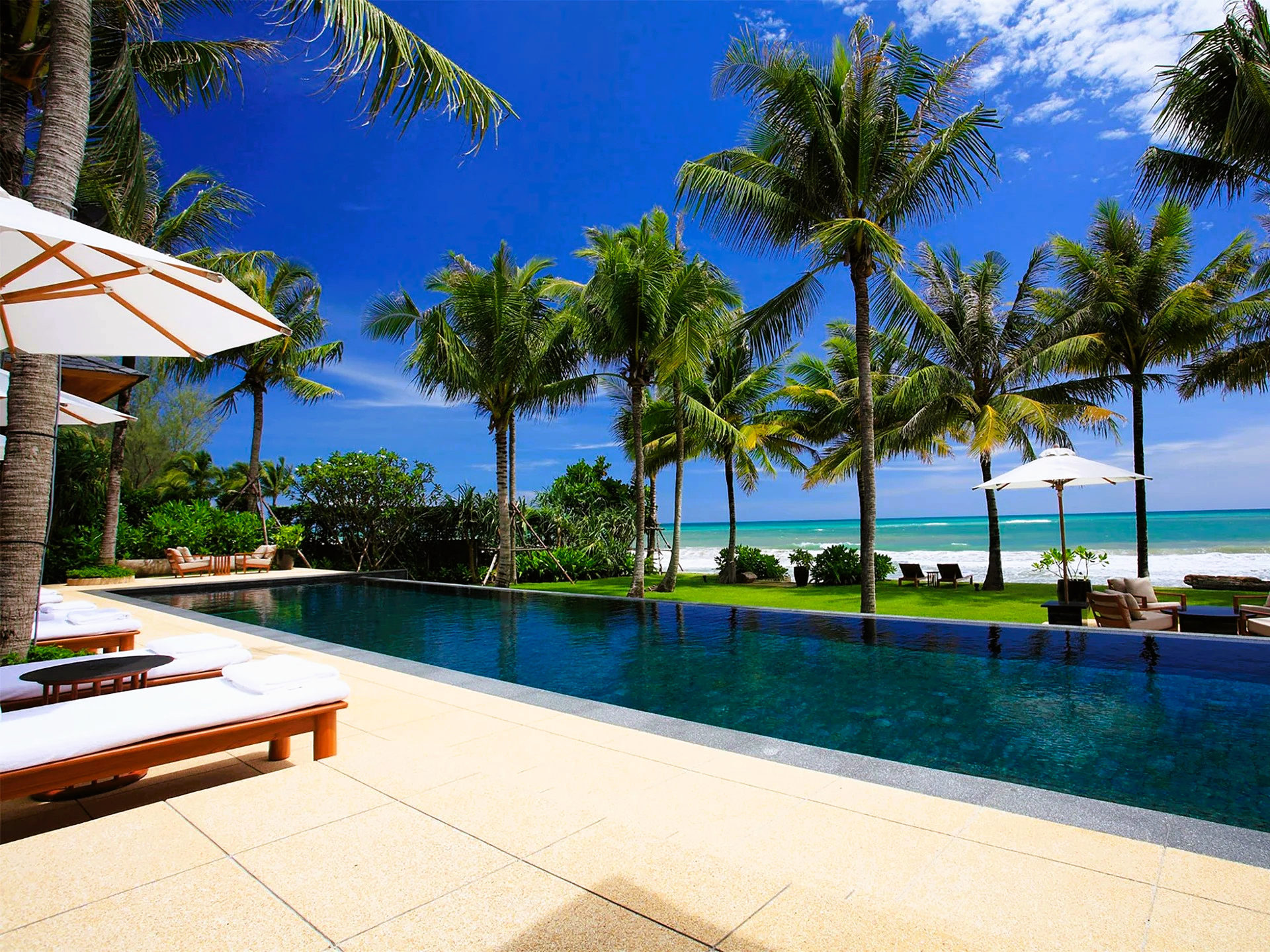 Poolside view of a resort with palm trees, ocean in the background, and sunny blue sky.