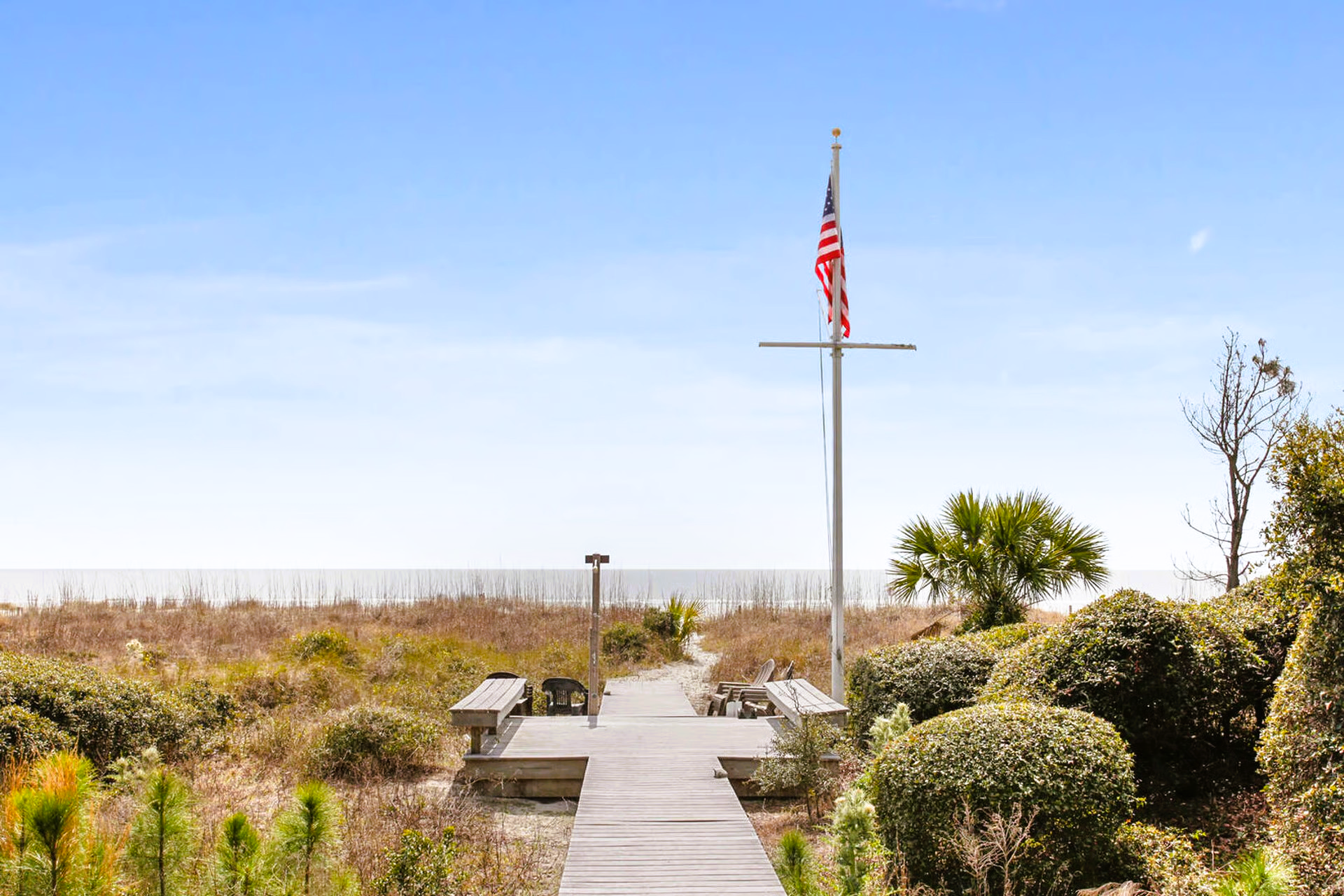 Wooden boardwalk leading to the beach, American flag, and greenery under a clear sky.