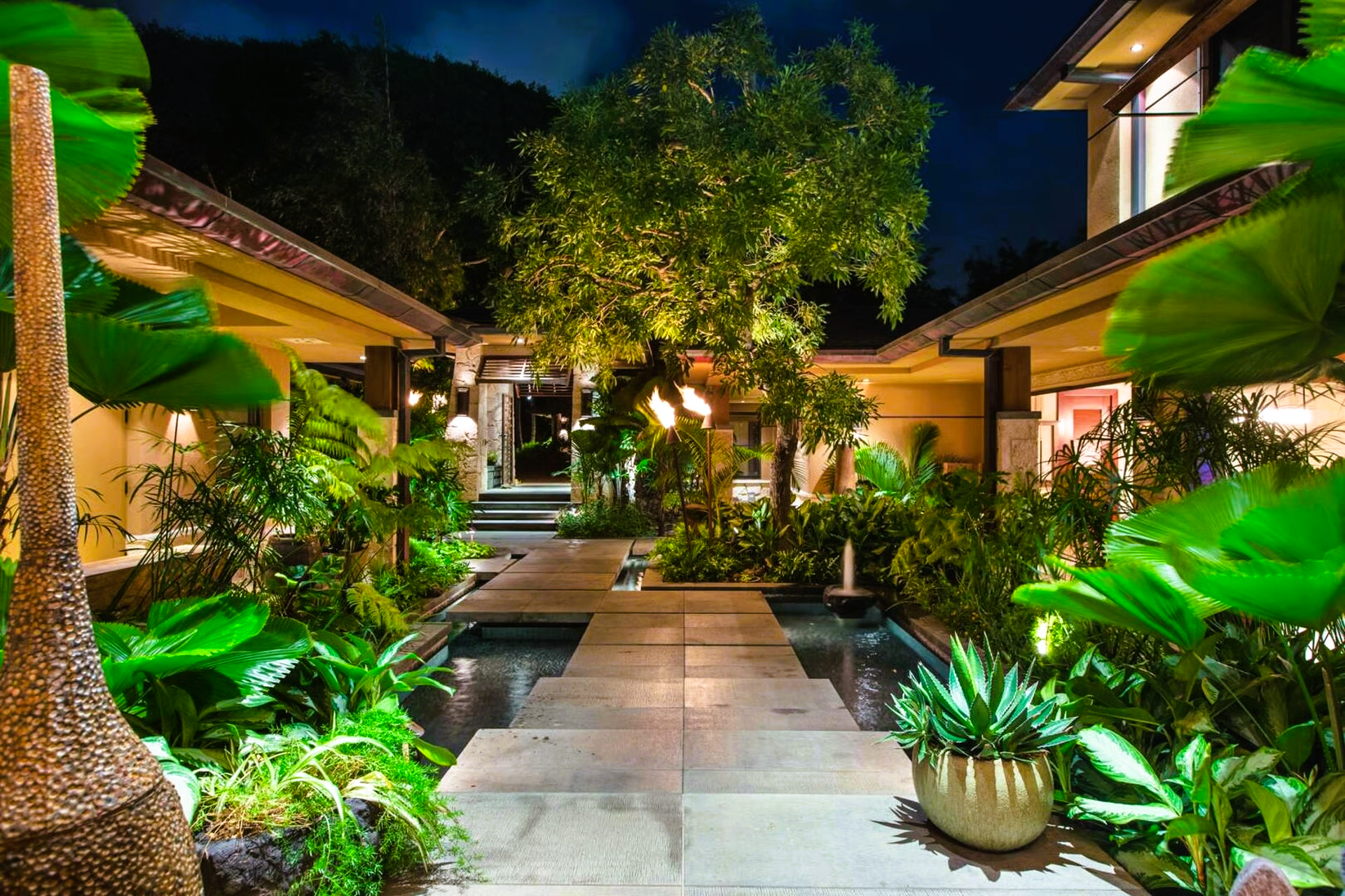 Stone path through a lush, illuminated tropical garden at night, leading to a building entrance.
