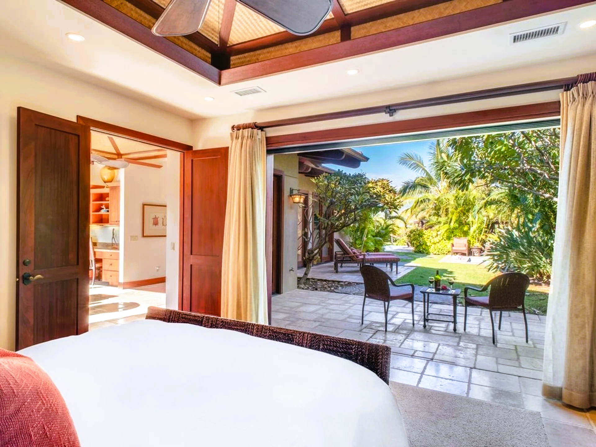 Bedroom with open doors to patio; bed in foreground, views of backyard, tropical plants.